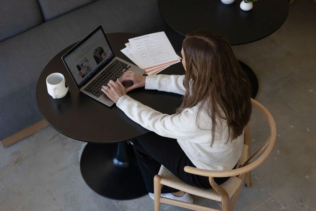 A woman with long brown hair sitting at a round black table working on a laptop with papers beside her, a white mug nearby, in a modern indoor setting.