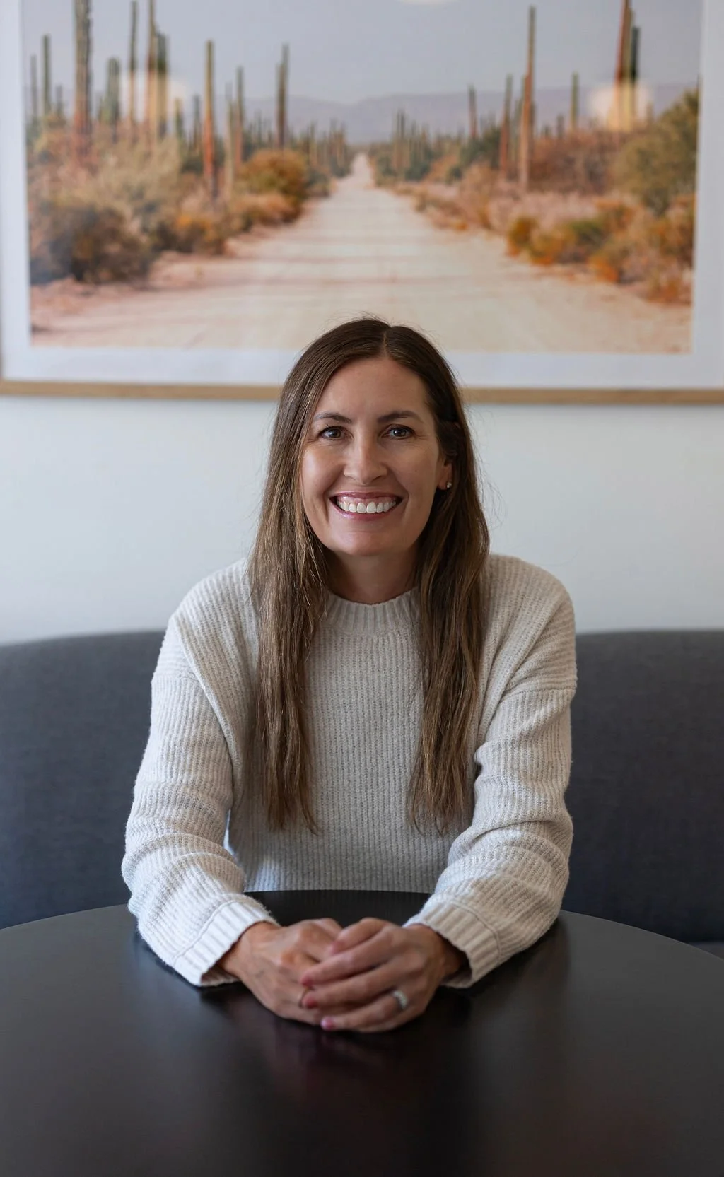 about- Jamie with long brown hair and a beige sweater is sitting at a black table, smiling. There is a sofa behind her and a large framed photo of a desert landscape with cacti in the background.