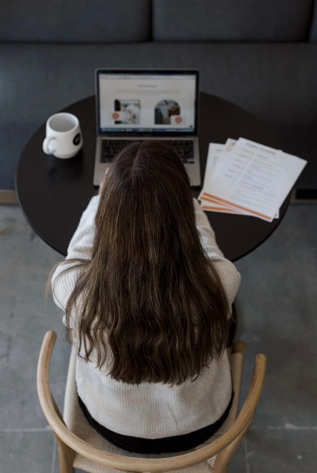 An overhead view of a woman with long brown hair sitting at a round black table with a laptop, a white coffee mug, and several documents spread out on the table.
