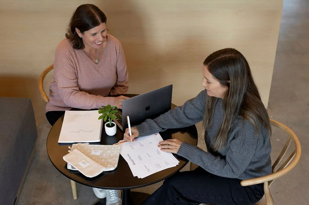 about Two women sitting at a round table with documents, a small potted plant, a laptop, and fabric samples, engaged in a discussion or meeting.