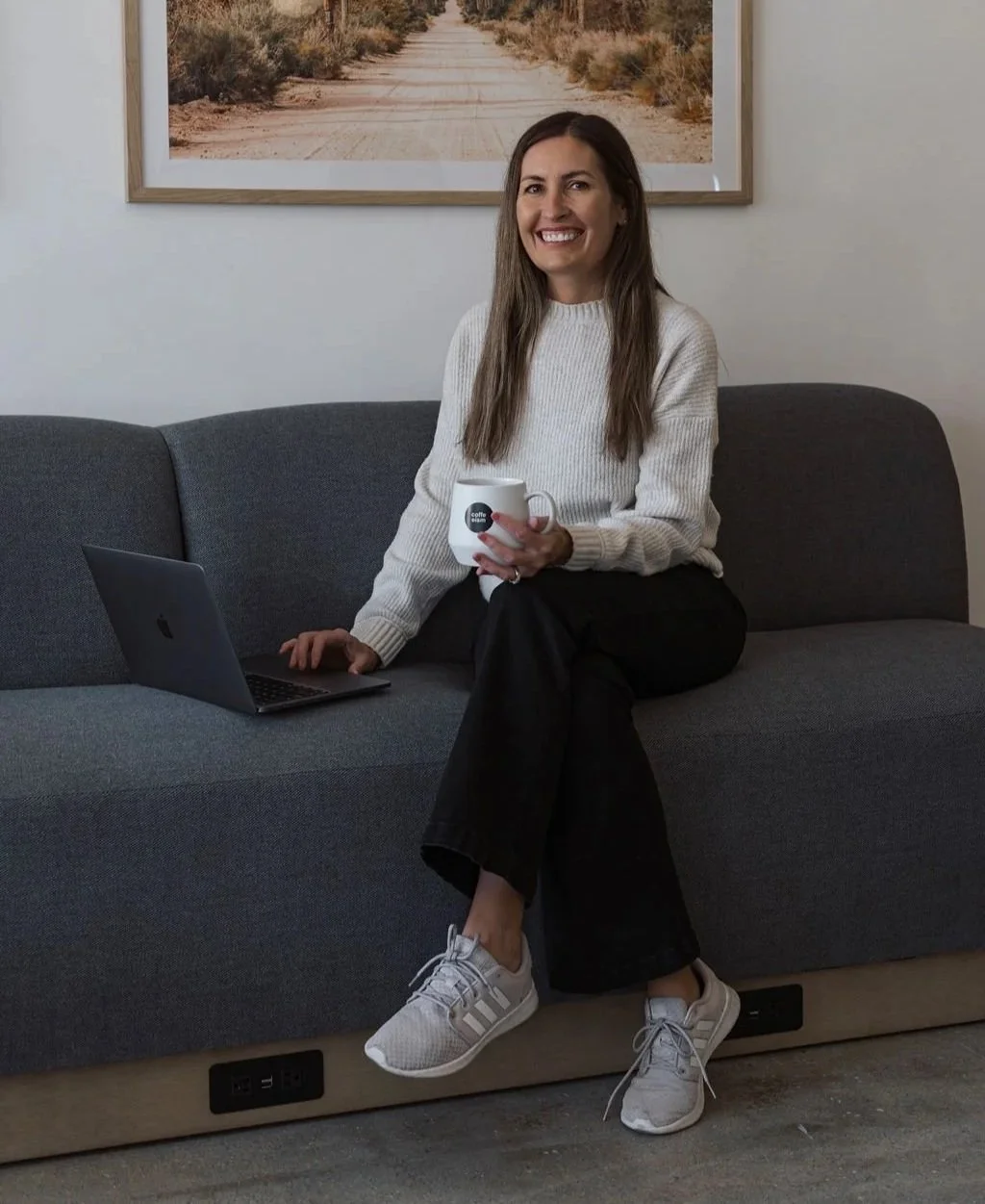 A Jamie sitting on a dark gray sofa, holding a white mug, with a laptop on the sofa beside her. She is smiling and wearing a white sweater, black pants, and gray sneakers. A framed picture of a desert road hangs on the white wall behind her.