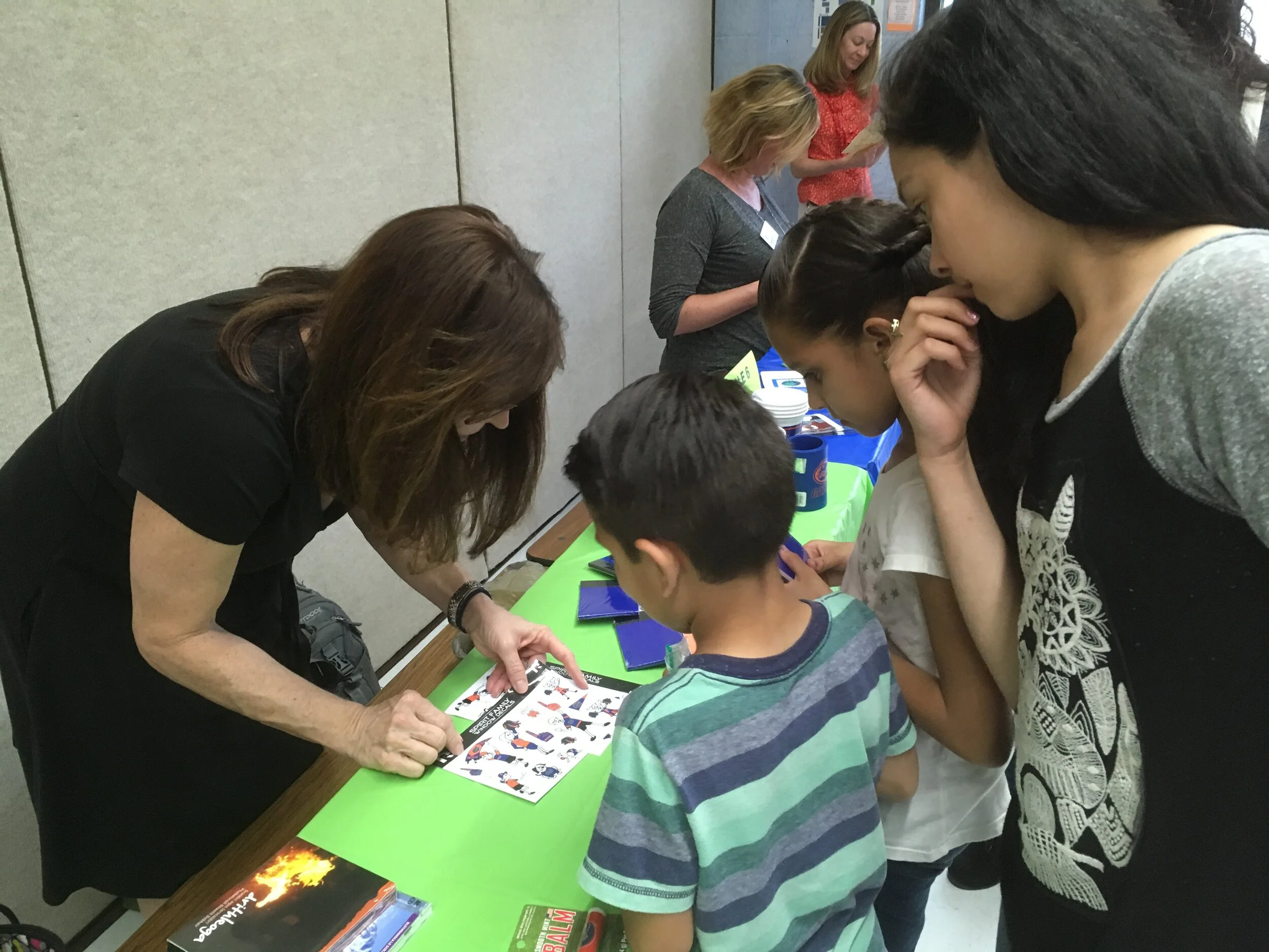 A woman showing a comic strip to two children, with other women in the background at a table with various items in a classroom or community center setting.