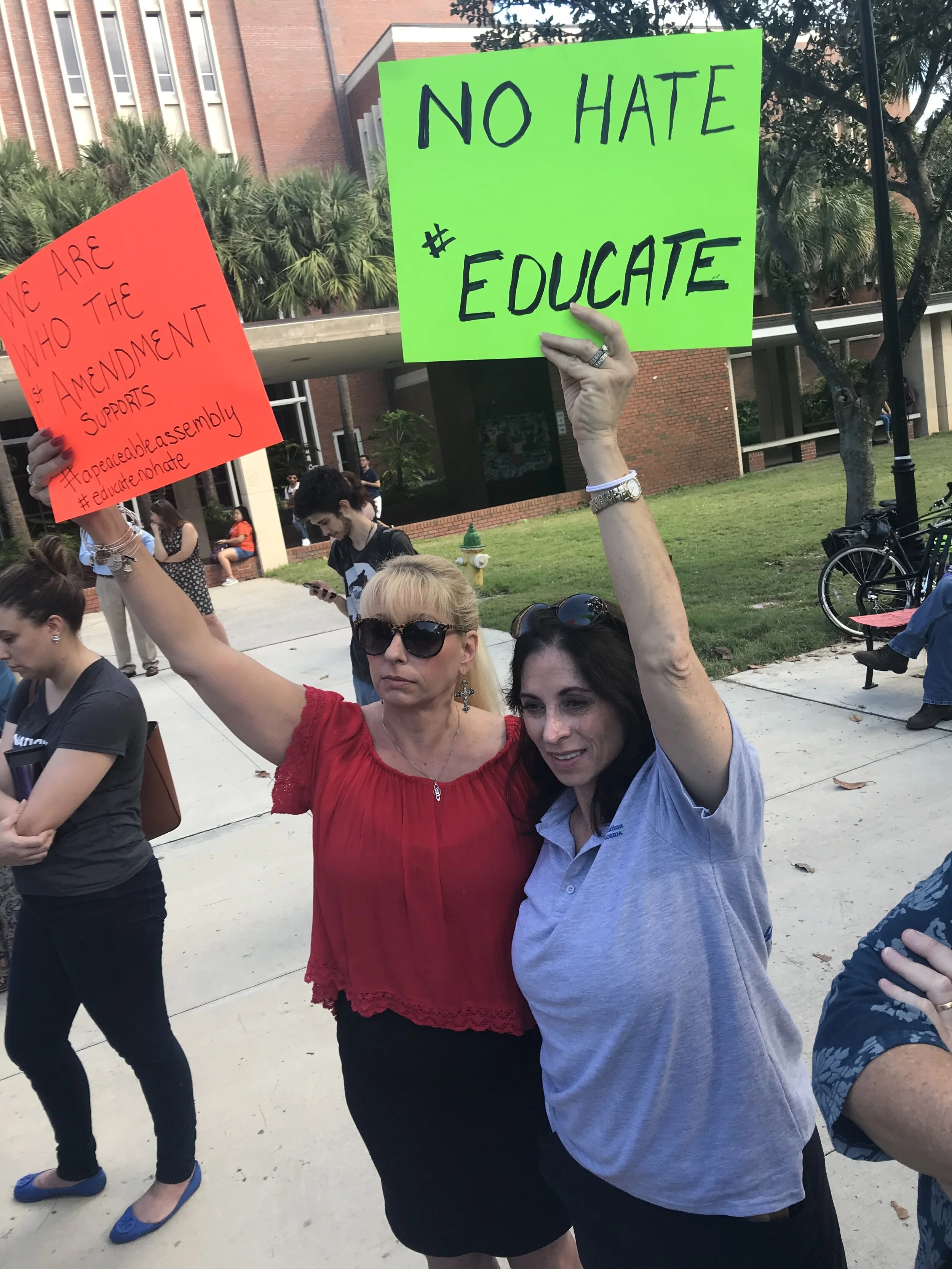 Two women holding protest signs outdoors during a demonstration; one sign reads 'NO HATE #EDUCATE' on a bright green poster, and the other red sign reads '#We are who the amendment supports #appeaceassembly #educateonhate'; people and a brick buildin