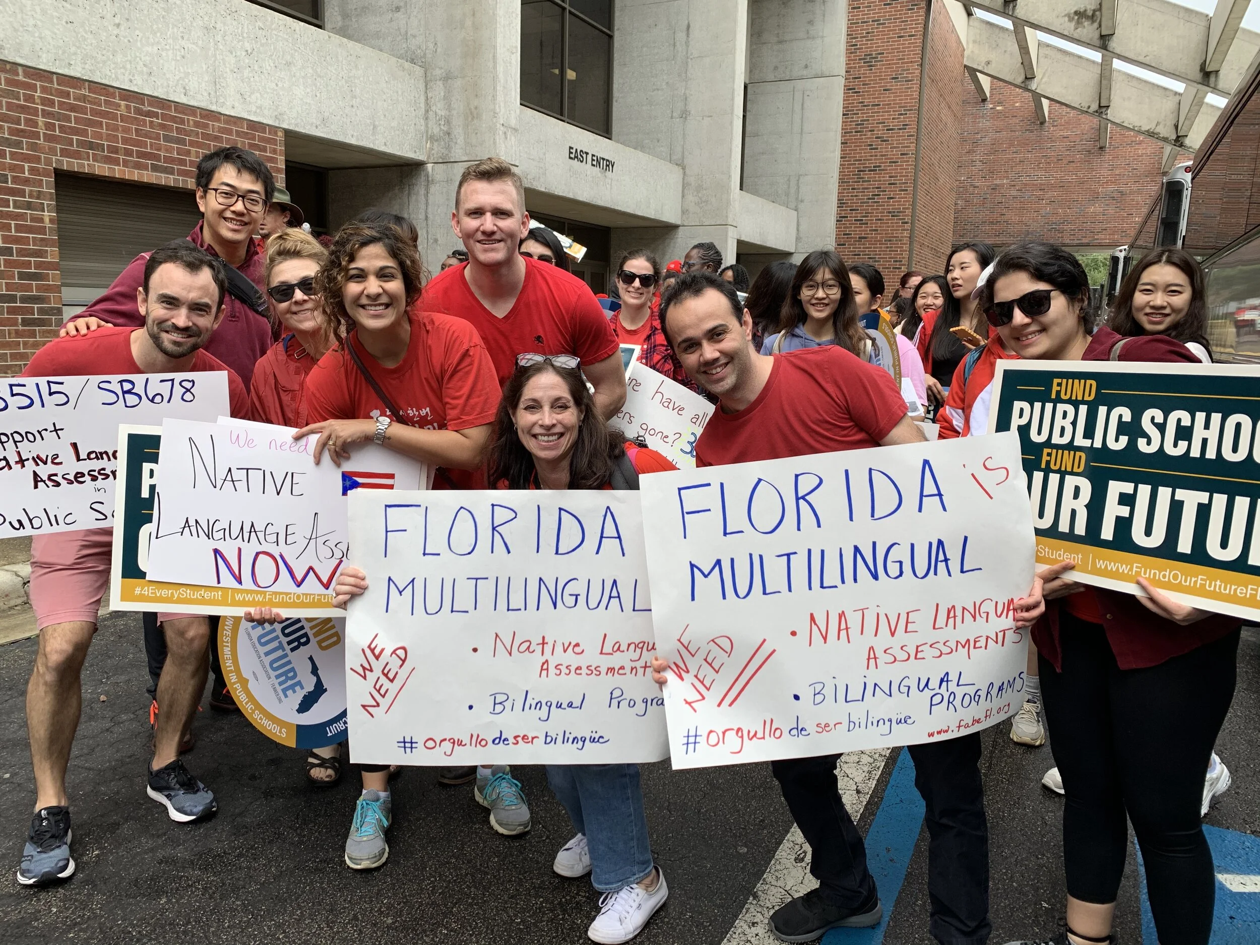 Group of people holding signs promoting bilingual programs and native language assessment outside a building.