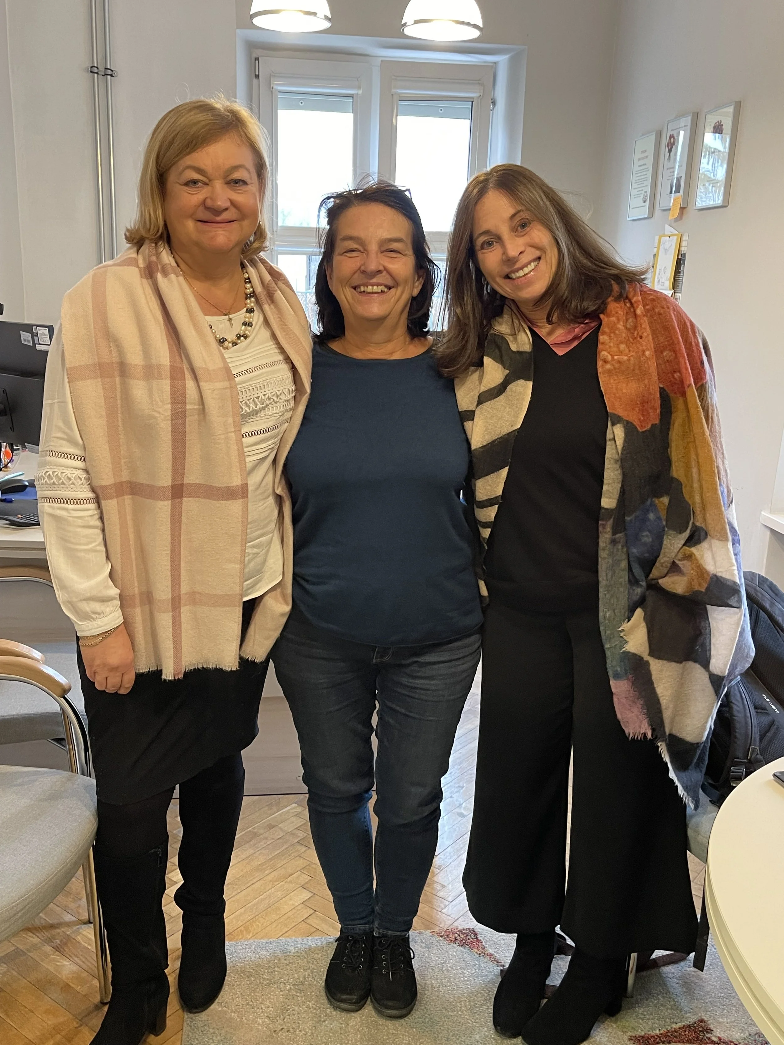 Three women standing together indoors, smiling for the camera, with a window behind them and framed pictures on the wall.