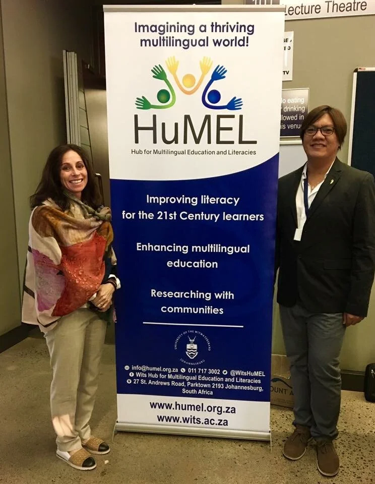 Two individuals standing next to a HuMEL banner at a conference or event. The banner promotes multilingual education and literacy improvements.