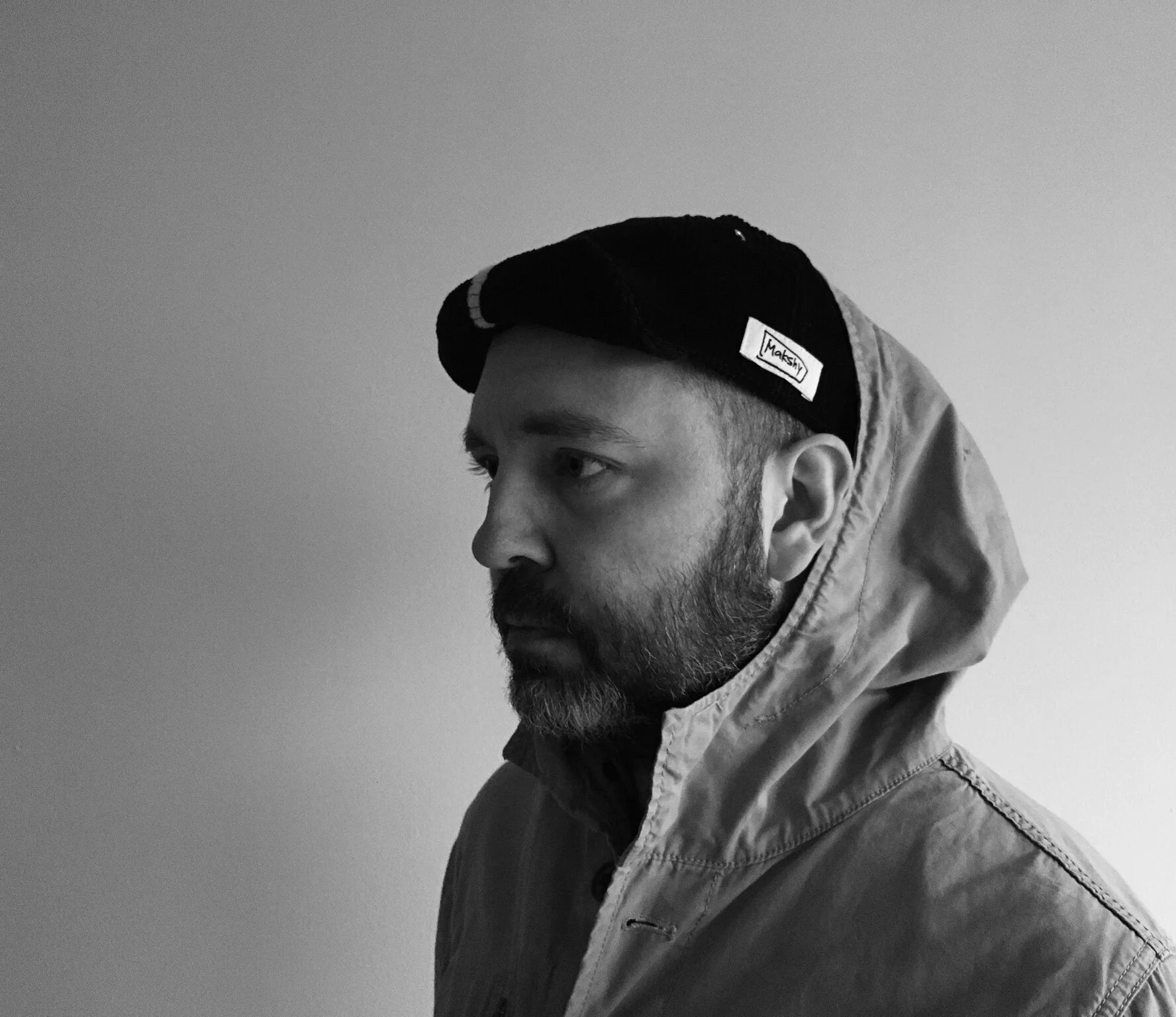 A man with a beard wearing a black, brimless Makshy docker cap, with the hood of his jacket up. The moody, black and white profile shot prominently features the white Makshy logo tag on the cap's cuff.

