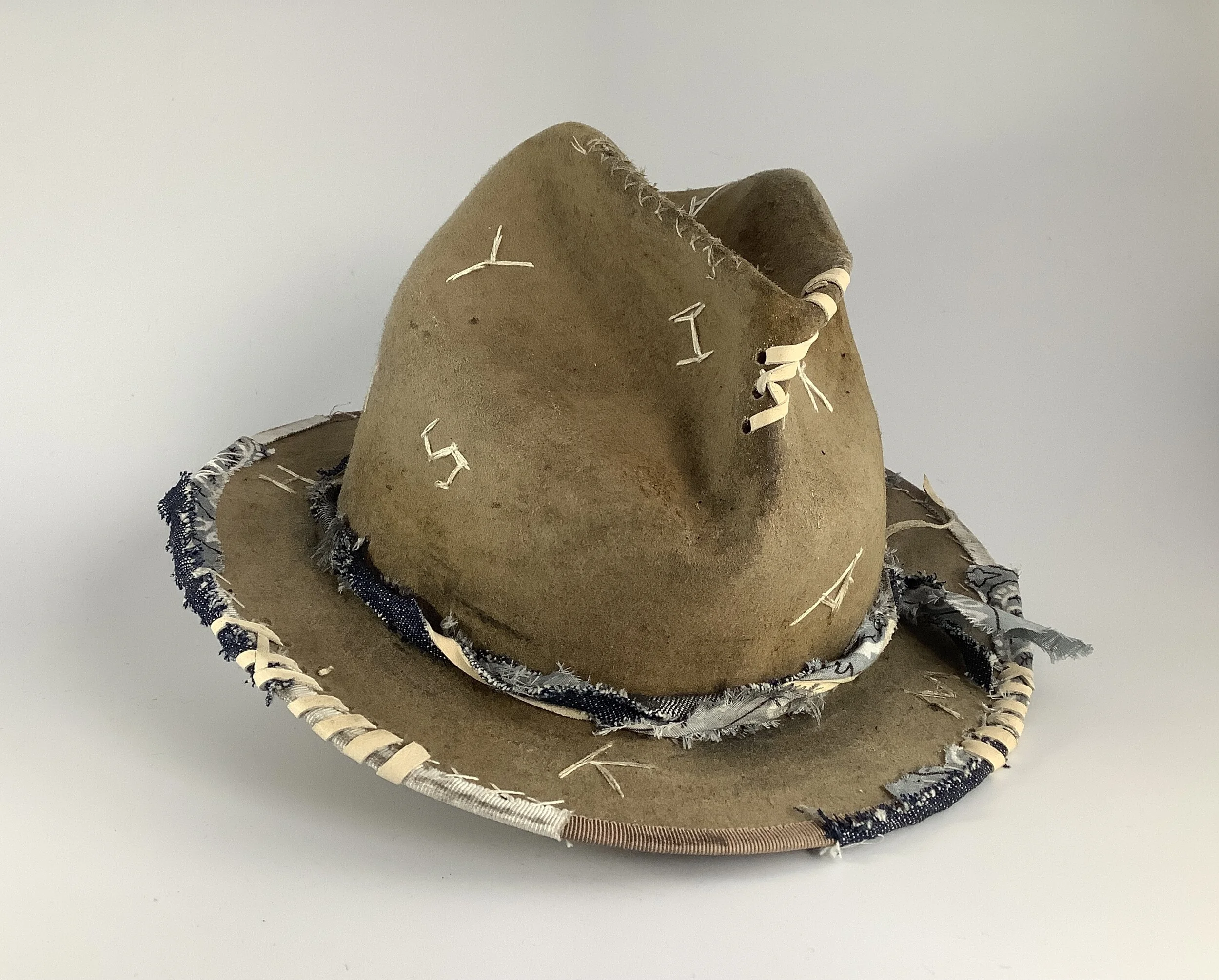 An example of wearable art from Makshy: a rustic, hand-aged brown fedora featuring primitive-style hand embroidery and a deconstructed brim bound with raw denim and fabric strips.