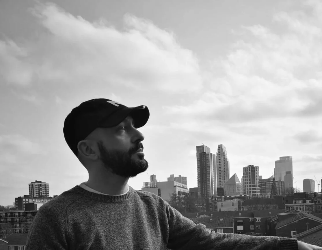 A black and white lifestyle shot of a man wearing a custom initial Makshy cap in an urban setting. He looks out over a view of city skyscrapers under a cloudy sky.