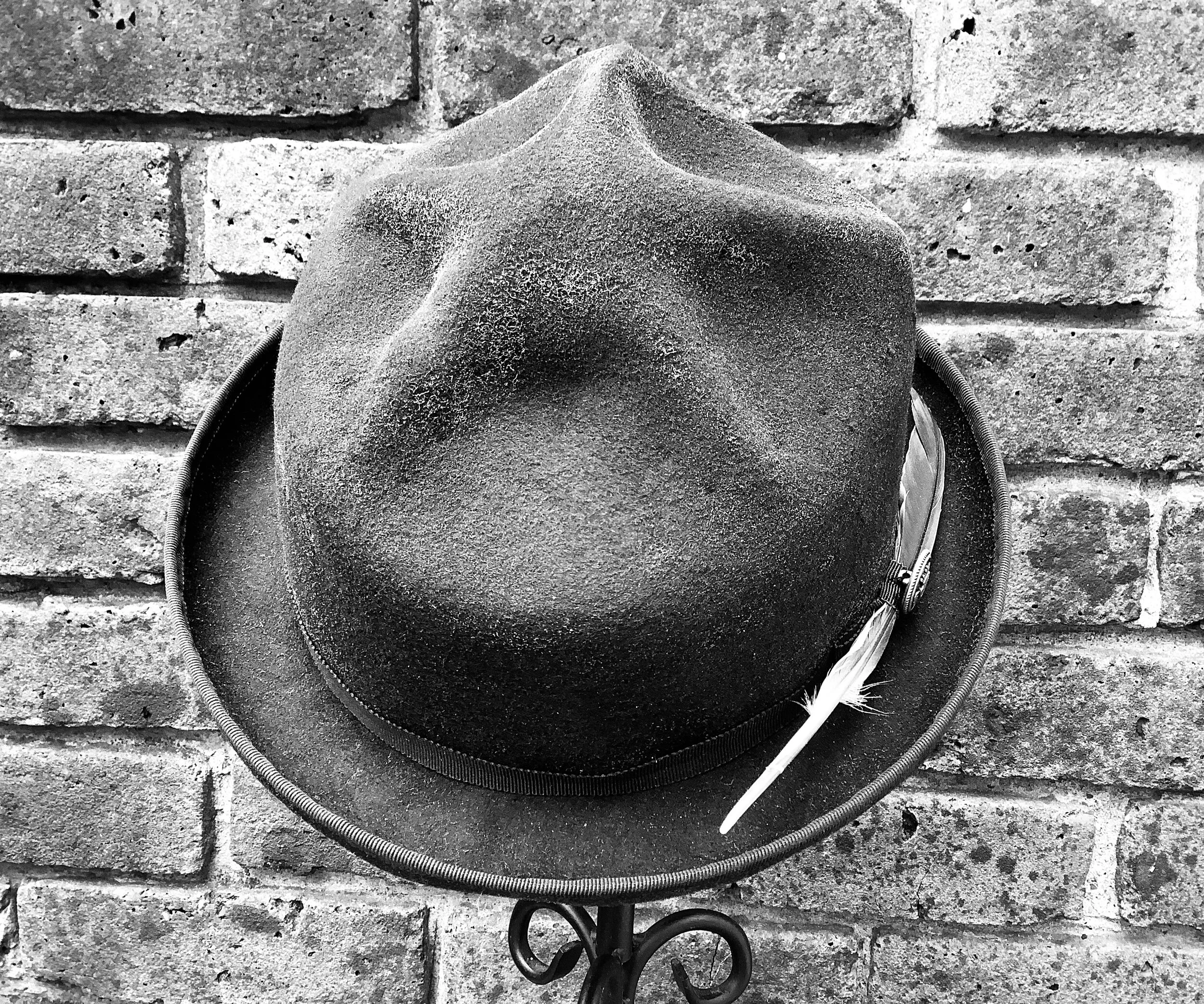 A Makshy Mountain Crown fedora, detailed with a long feather, is displayed on an ornate metal stand. The high-contrast black and white photo is set against a rustic, weathered brick wall, emphasizing the textures.