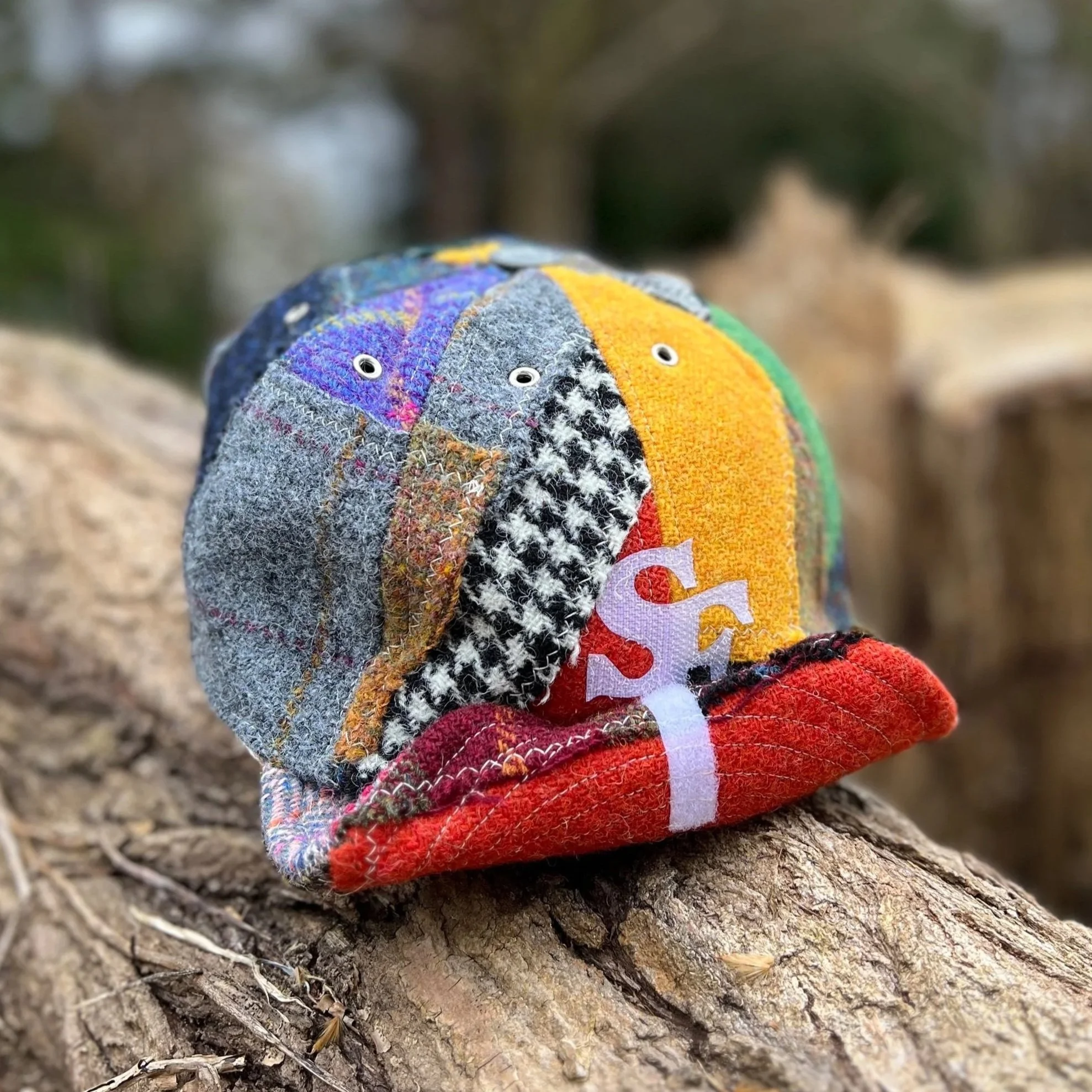 The rich textures of a bespoke Makshy Millinery cap, showcased against the natural bark of a log. The cap features a colorful patchwork of tweeds, a bold houndstooth section, and is personalized with the initial 'S'.