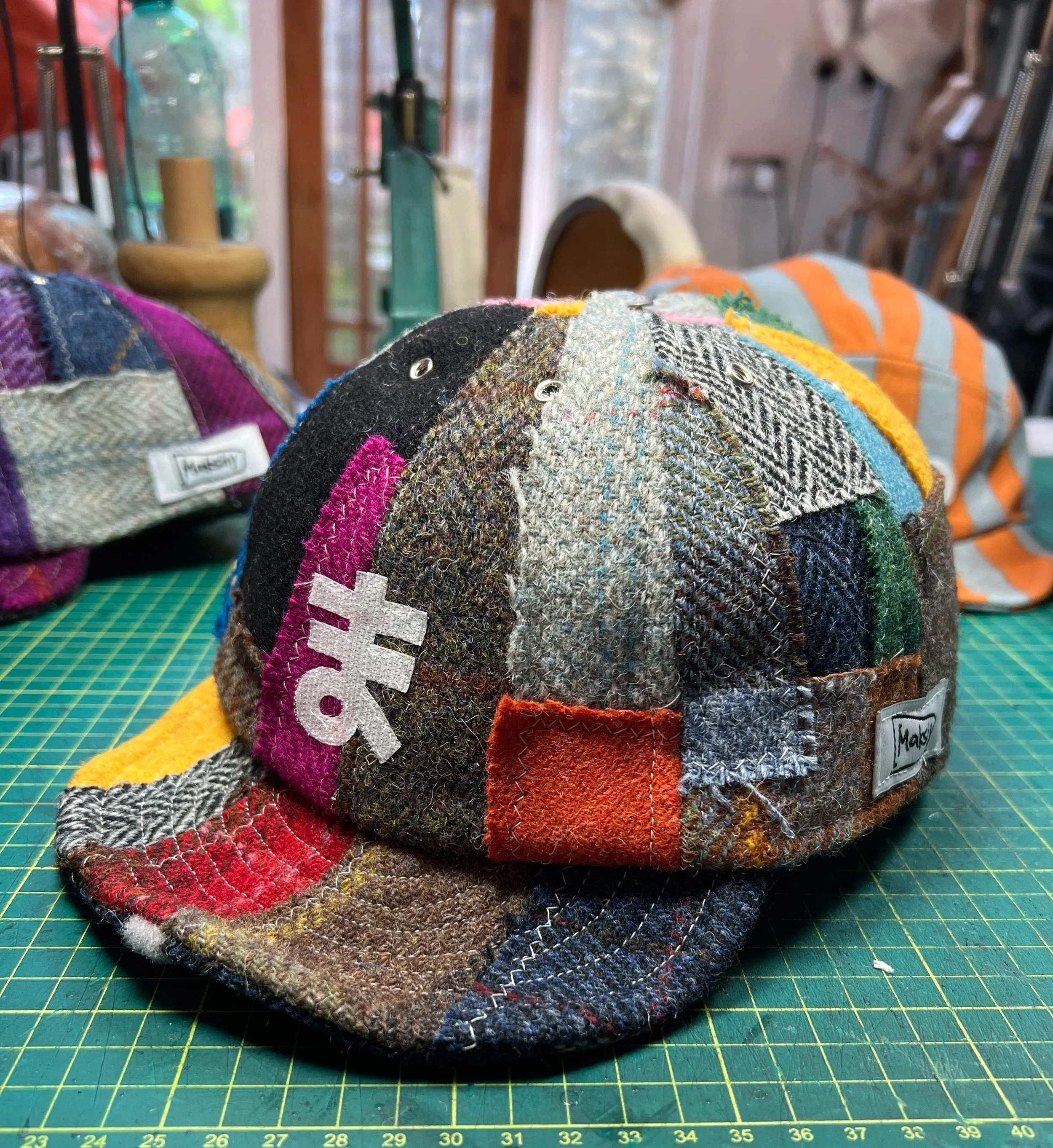 A vibrant, multi-colored patchwork Harris tweed cap by Makshy, sitting on a cutting mat in the brand's workshop. The cap is customized with the white Japanese Katakana character 'Ma' on the front, reflecting the brand's name.