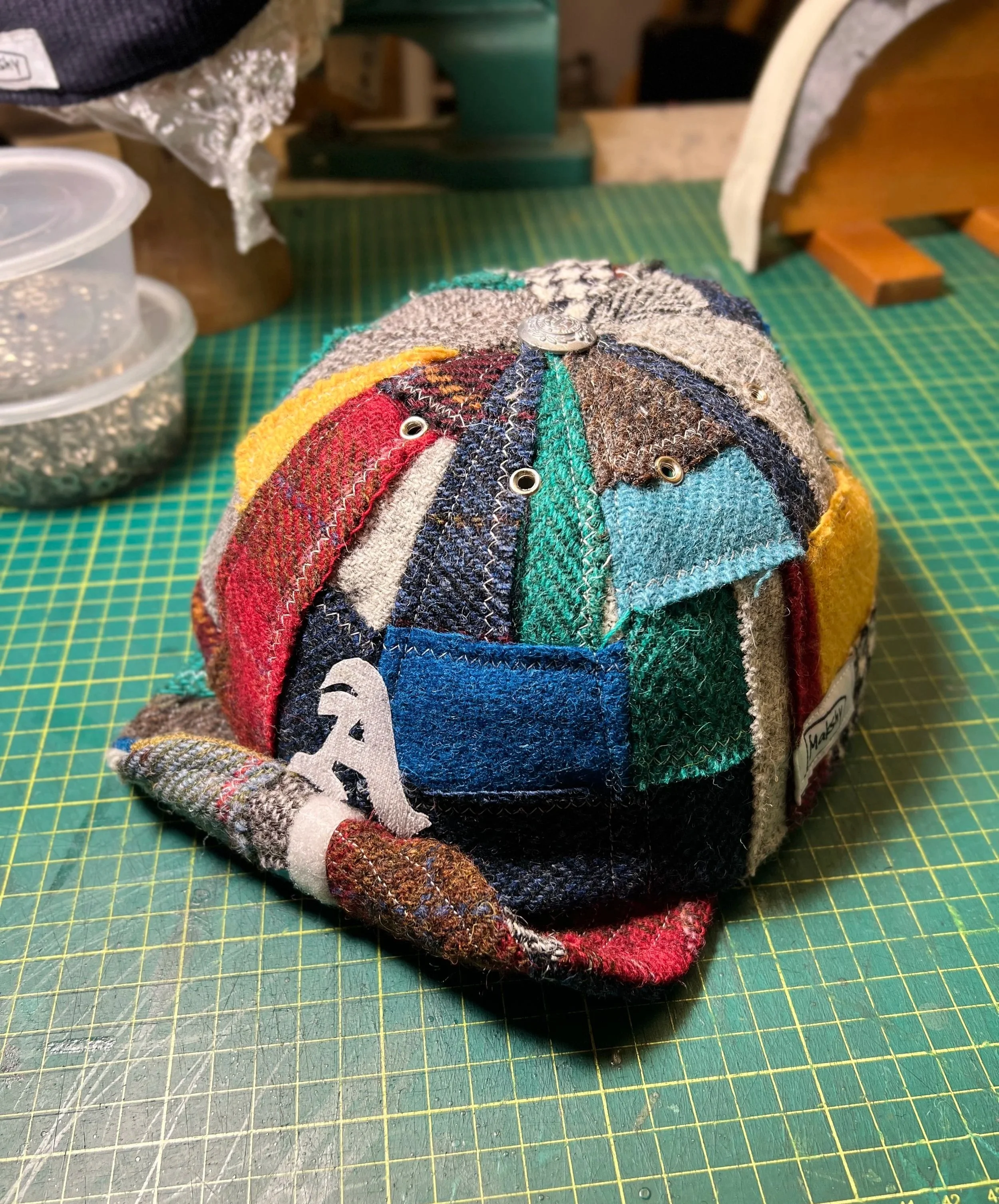 A vibrant, multi-colored patchwork Harris tweed cap by Makshy, sitting on a cutting mat in the brand's workshop. This custom piece features a white cursive initial 'A' on a blue patch at the front.