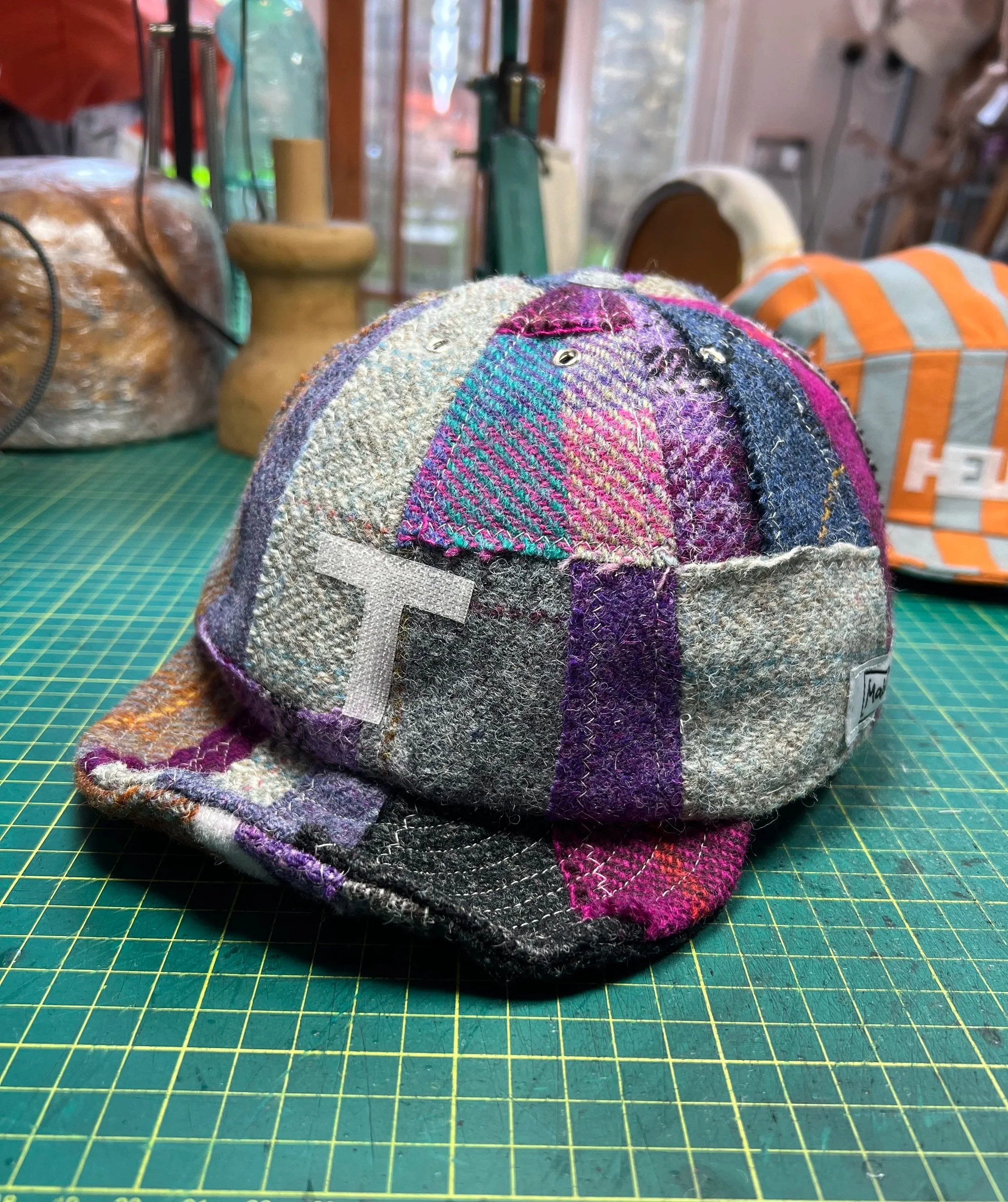 A bespoke patchwork tweed cap by Makshy Millinery, displayed on the workbench where it was made. This colorful version, featuring the initial 'T', showcases a vibrant mix of purple and grey wools.