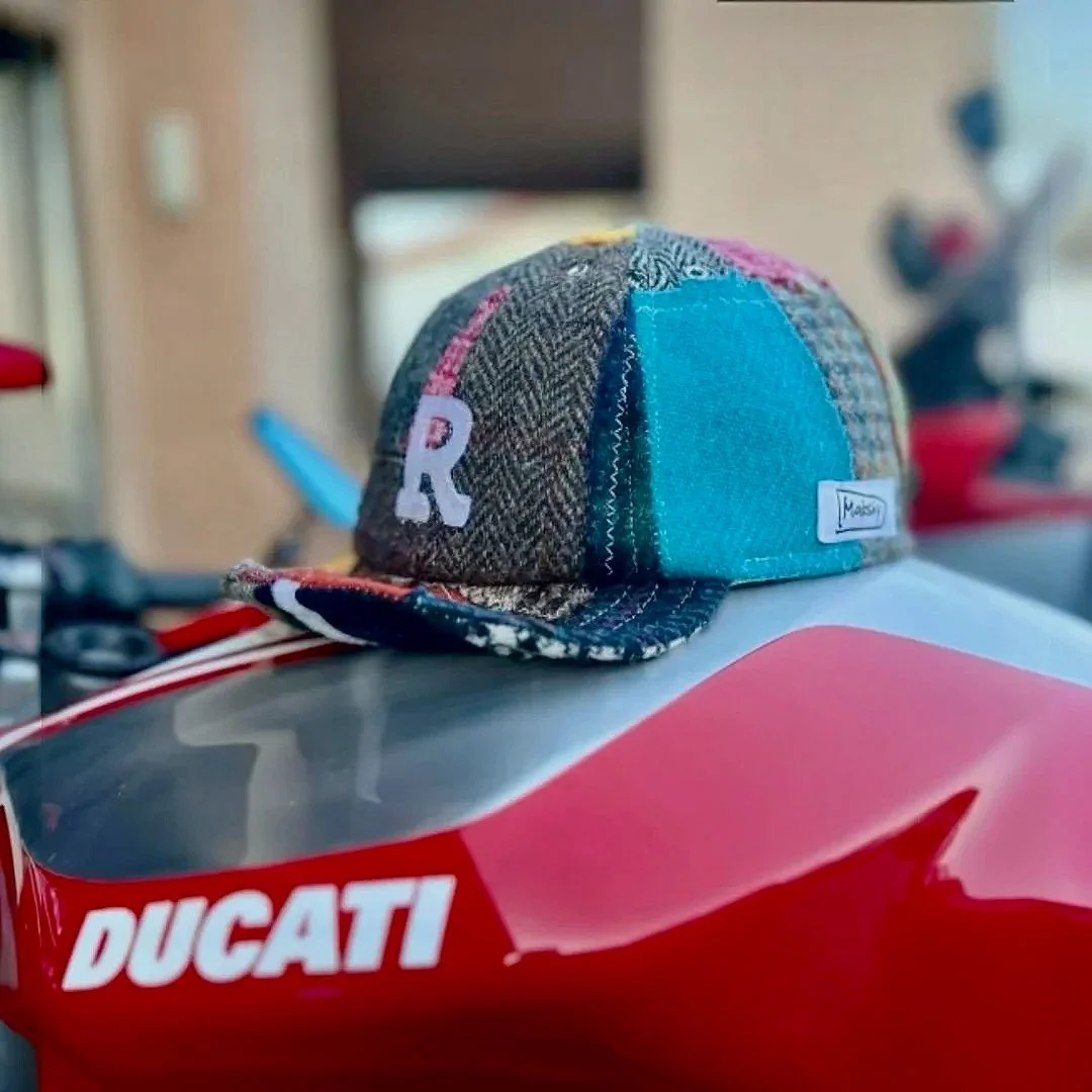 Showcasing the versatile style of Makshy Millinery, a colorful, handmade patchwork cap is displayed on a high-performance Ducati motorcycle. The artisanal tweed cap contrasts with the bike's modern design.