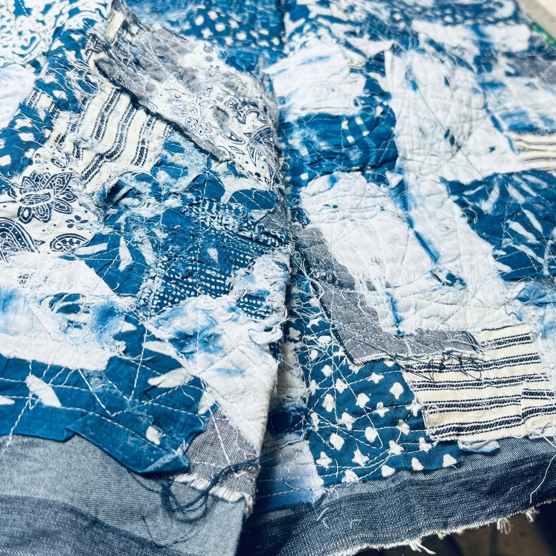 A close-up of a textural, handcrafted fabric from Makshy Millinery, featuring a patchwork of blue and white textiles in various patterns like paisley, shibori, and stripes. The piece is detailed with frayed edges and visible sashiko-style quilting.