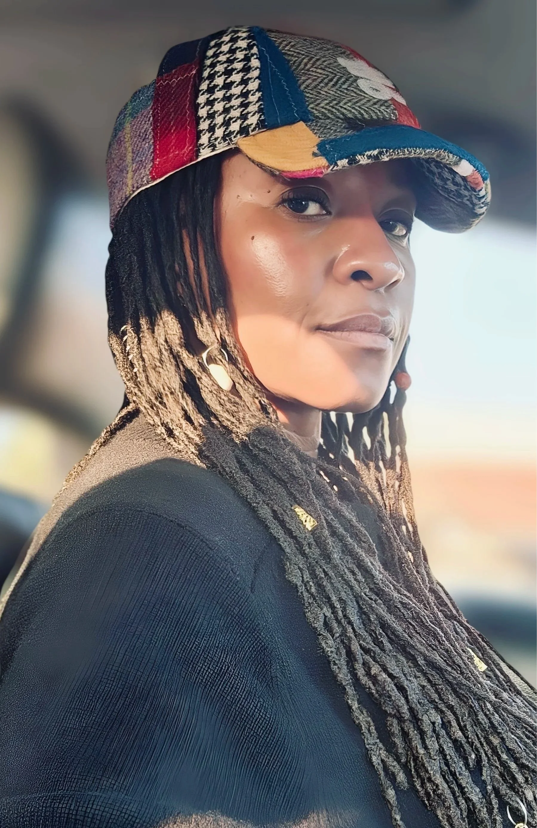 A confident woman with long locs, looking over her shoulder while wearing a colorful Makshy patchwork tweed cap. The cap features a houndstooth brim, and she is bathed in warm, golden sunlight.