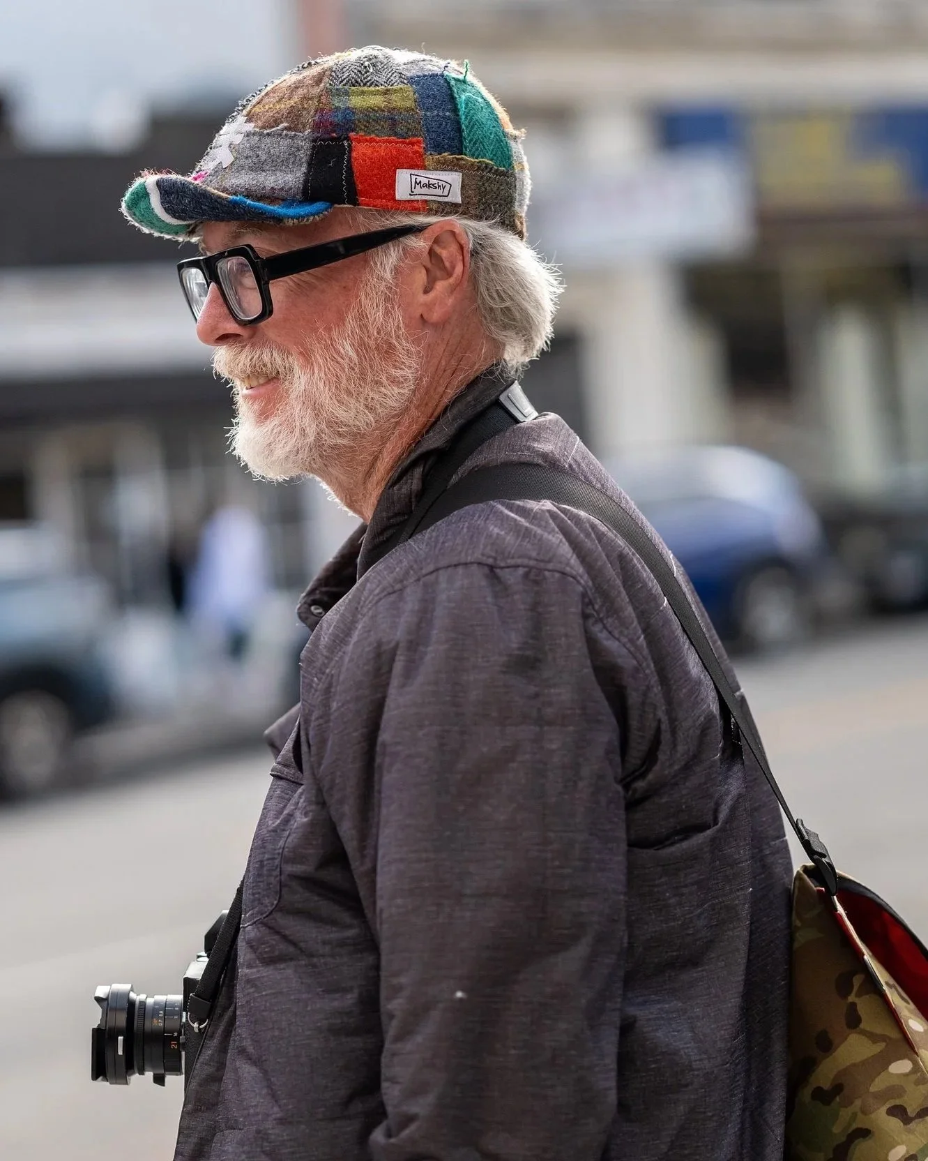 A candid street-style portrait of a man in glasses wearing a colourful patchwork Harris Tweed cap by Makshy. The cap’s vibrant mix of textures and tones adds character to his casual look as he smiles against an urban backdrop with a camera slung over