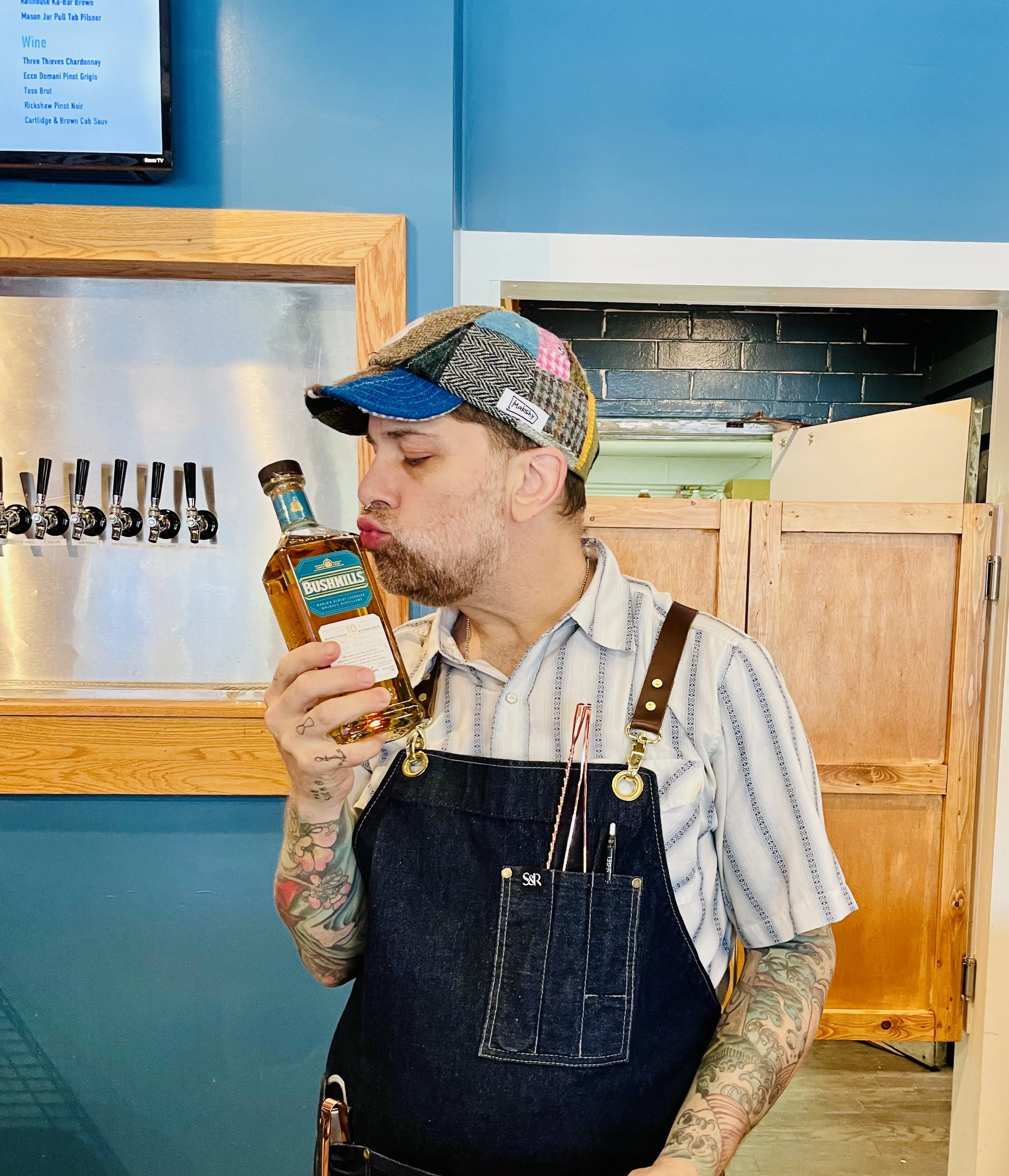 A bartender wearing a colorful Makshy patchwork tweed cap playfully kisses a bottle of Bushmills whiskey. He is standing behind the bar, wearing a denim apron, with beer taps visible in the background.