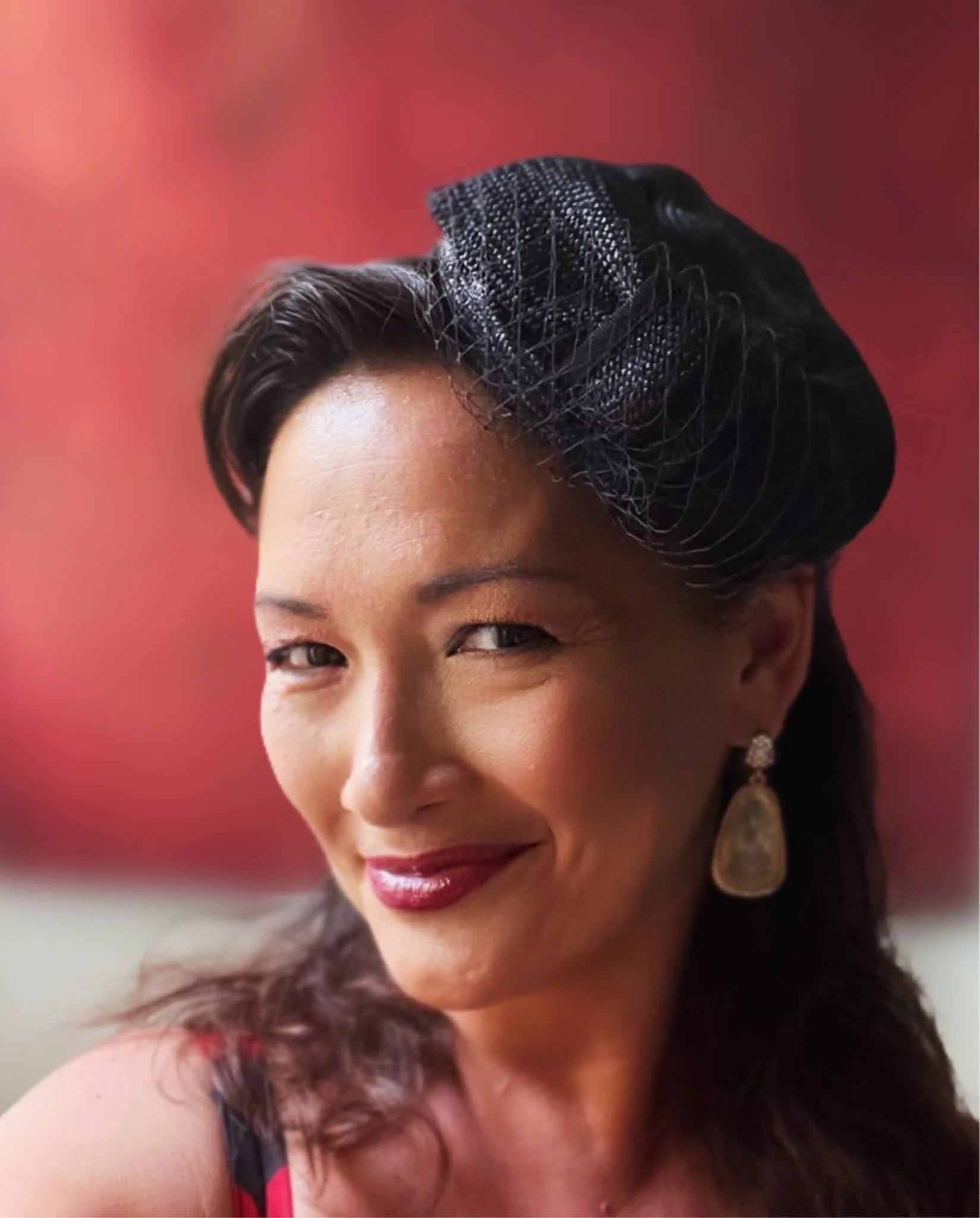 An elegant woman with dark hair and red lipstick smiles at the camera while wearing a black Makshy fascinator. The woven straw-style hat is tilted on her head and features a delicate birdcage veil. The portrait has a warm, red background.