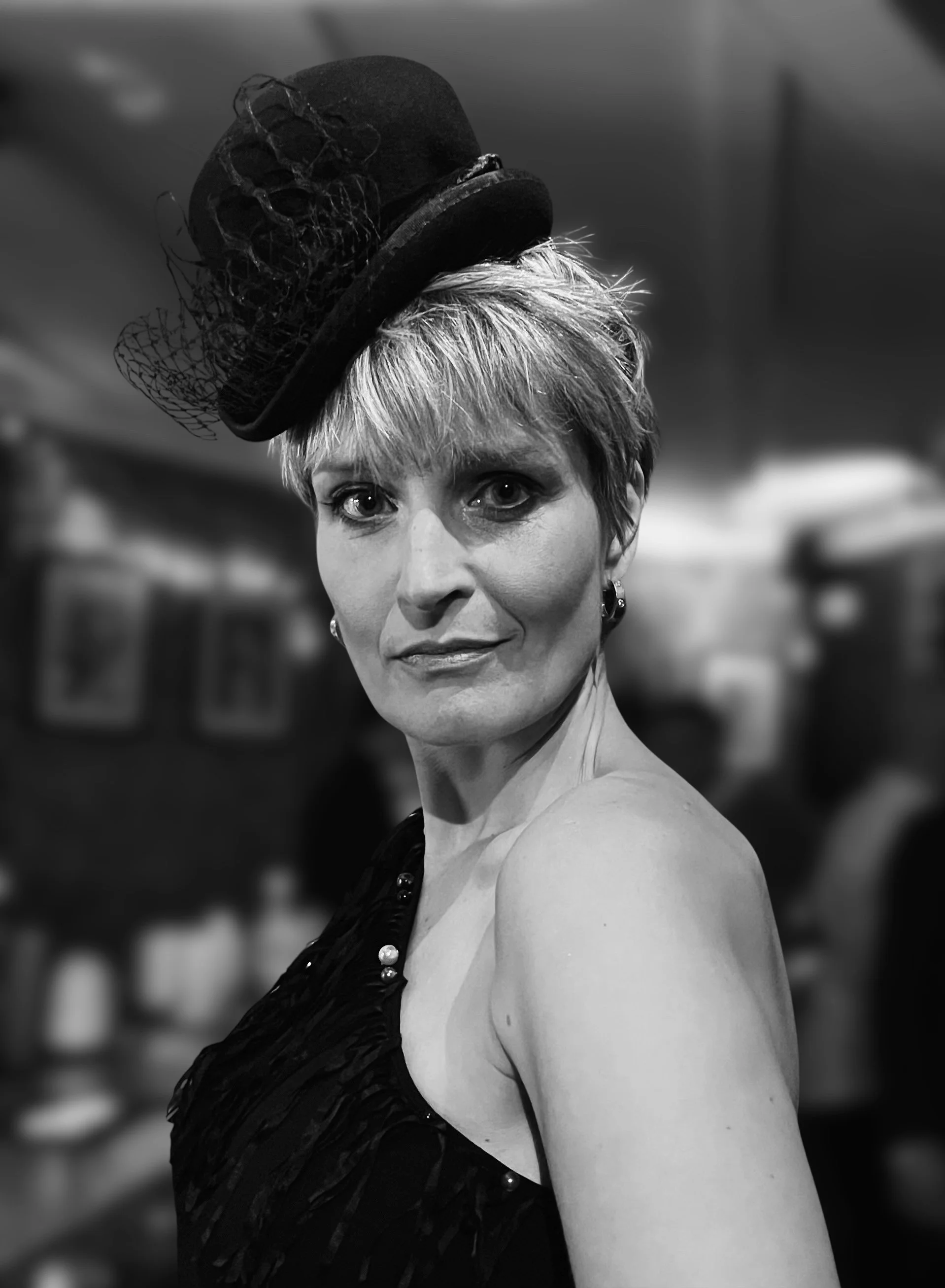 A glamorous, black and white photo of a woman modeling a statement piece from Makshy Millinery. Her tilted mini top hat, adorned with feathers and a veil, perfectly complements her confident gaze and one-shoulder dress.