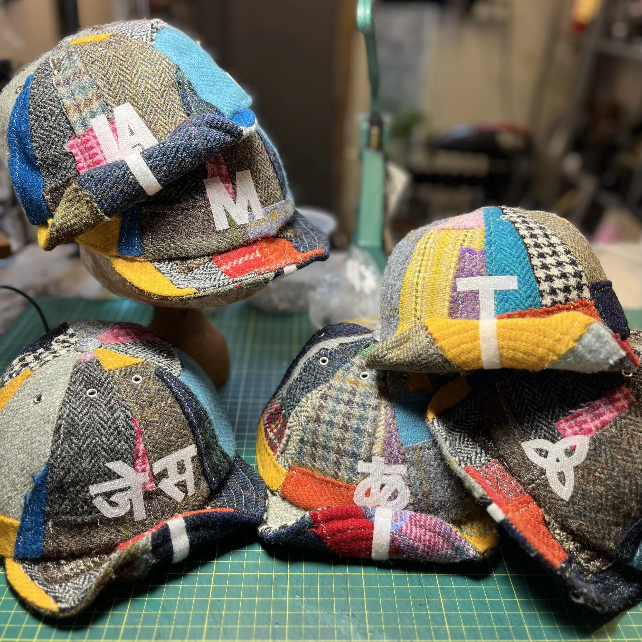 A collection of four unique, custom-made patchwork tweed caps from Makshy, displayed in the workshop. The image showcases a variety of personalized motifs, including the words "I AM," characters in Devanagari script, a Japanese character, and a Celti