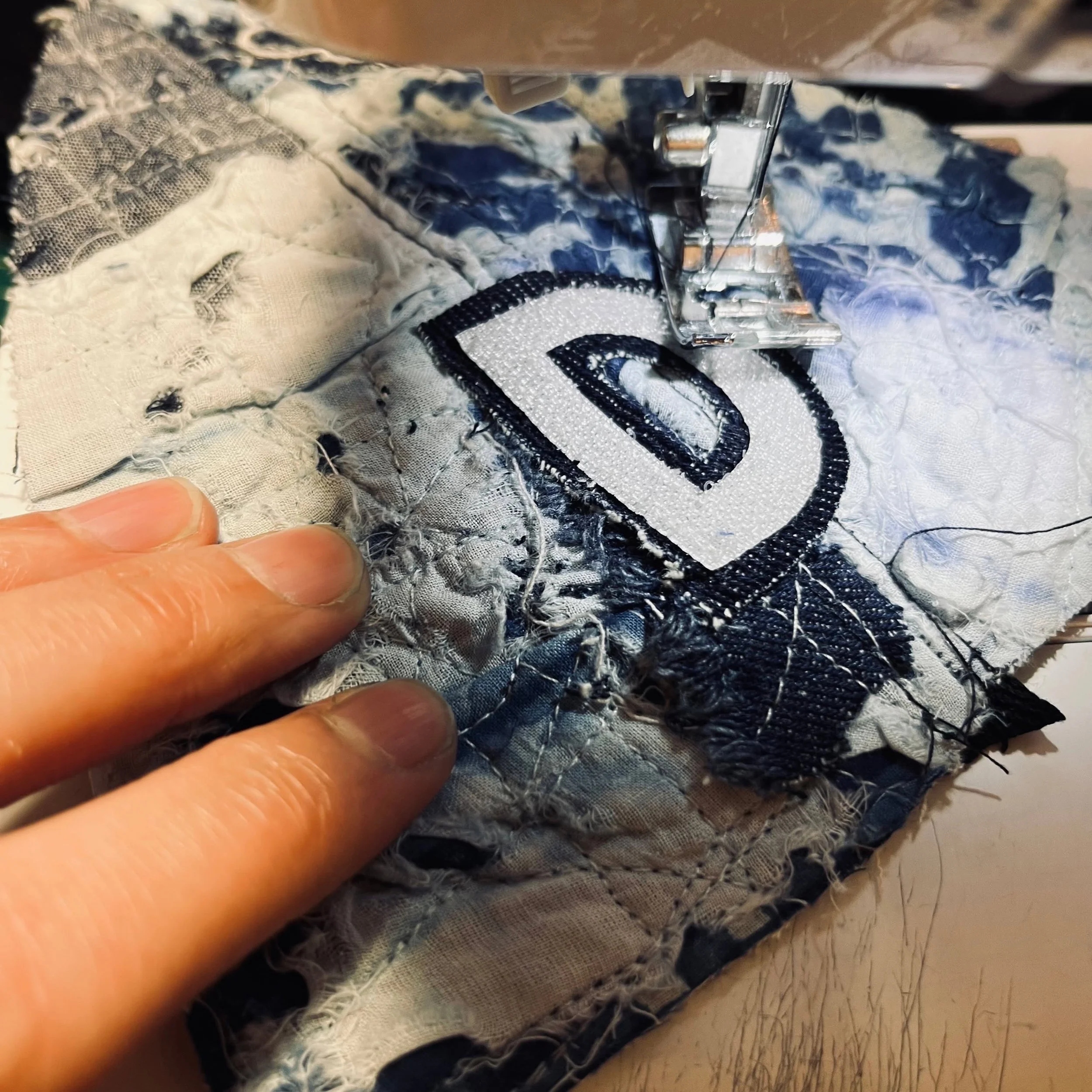 The hands-on crafting process at Makshy Millinery. A milliner's hand guides a piece of blue and white Boro-style patchwork fabric as a sewing machine stitches on a white letter 'D' appliqué.