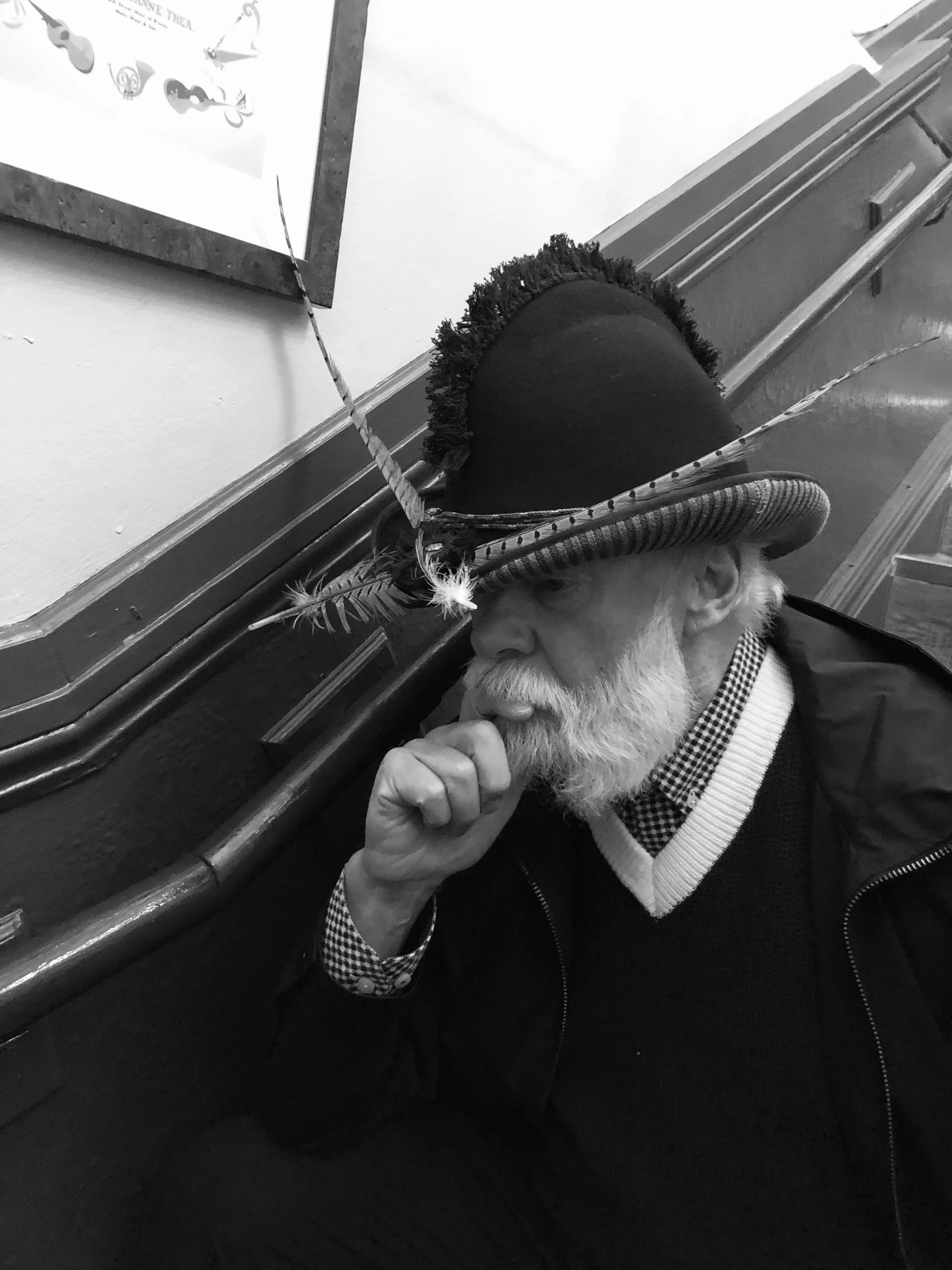 A distinguished older gentleman with a white beard, wearing a unique Makshy hat in a thoughtful pose. The hat features a dramatic, brush-like crest over the crown and is decorated with several long feathers. The portrait is in black and white.


