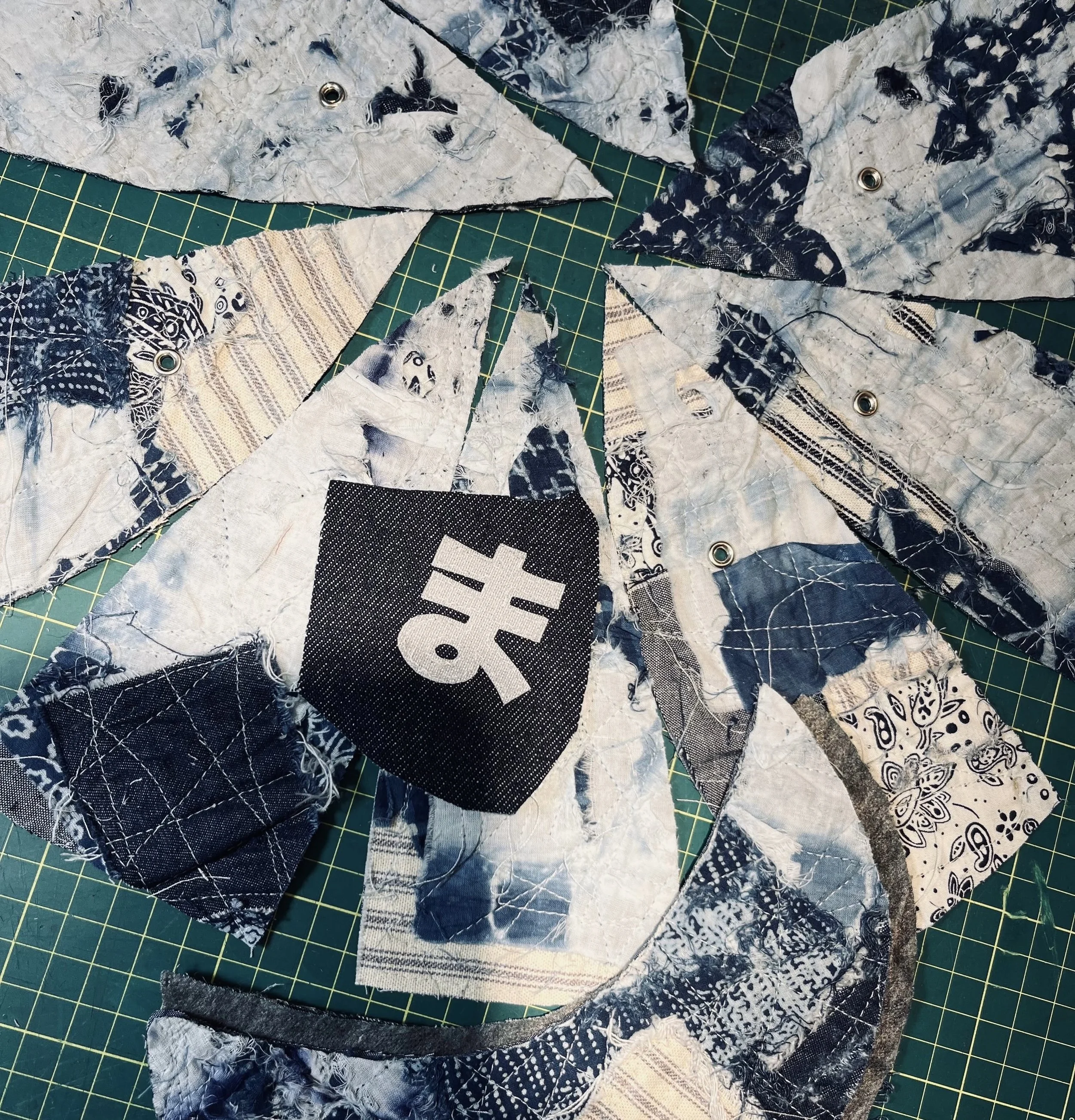 A collection of handcrafted panels for a Makshy Boro cap, arranged in a circle on a green cutting mat. The patchwork panels are made of indigo and white fabrics, with a central black patch featuring the Japanese hiragana character 'ma'.