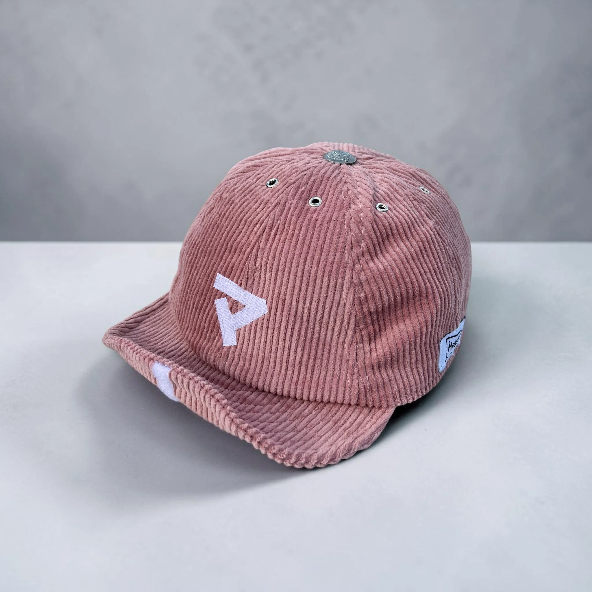 Pink corduroy 8 panel baseball cap with a structured crown, metal eyelets, and a shapeable roll-up visor, featuring a white katakana マ (ma) motif on the side, displayed on a neutral grey background.