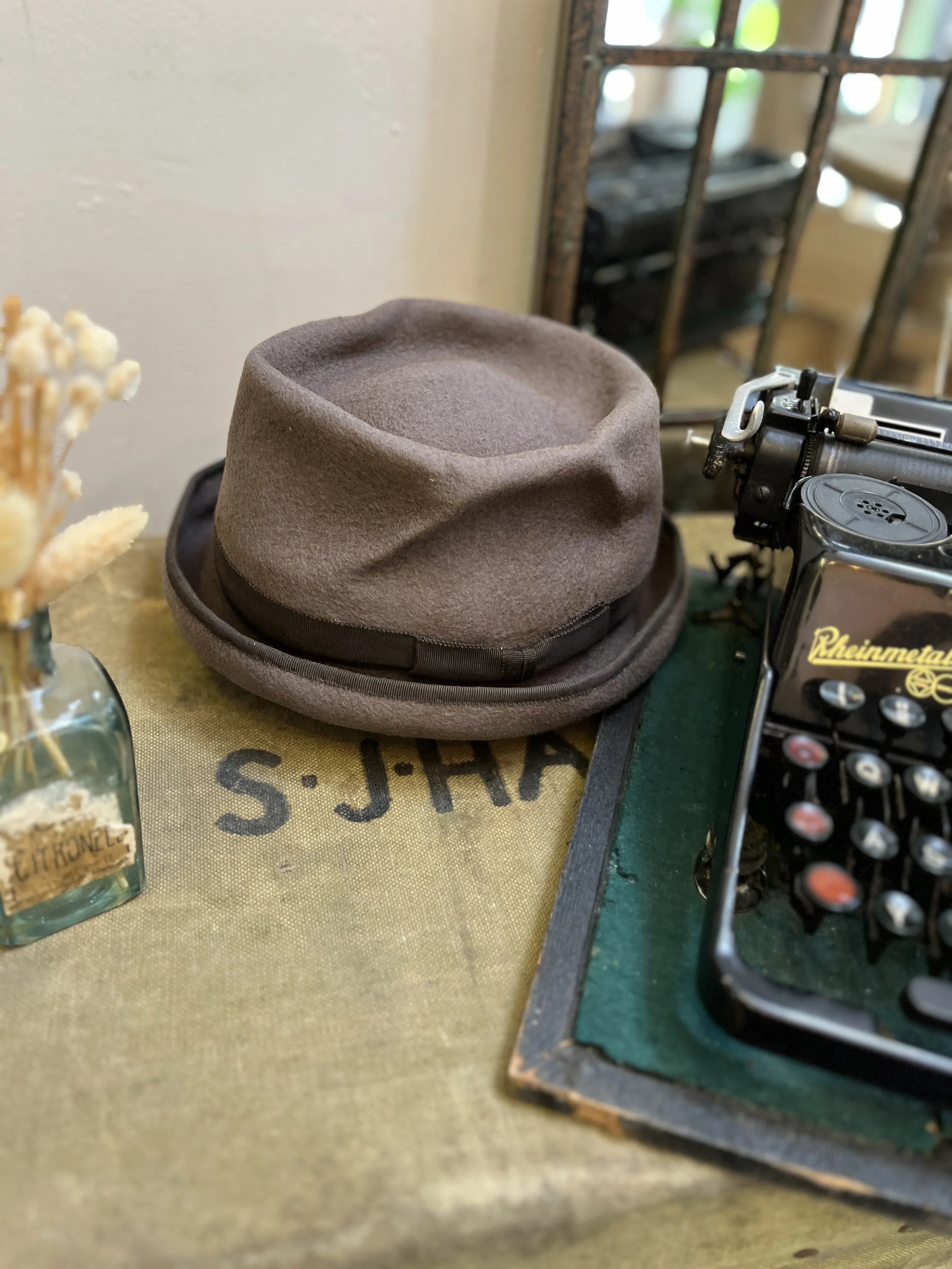 An atmospheric photo that tells a story of classic style, featuring a Makshy fedora alongside a vintage typewriter. The warm, nostalgic scene is completed with dried flowers and a rustic canvas cloth.