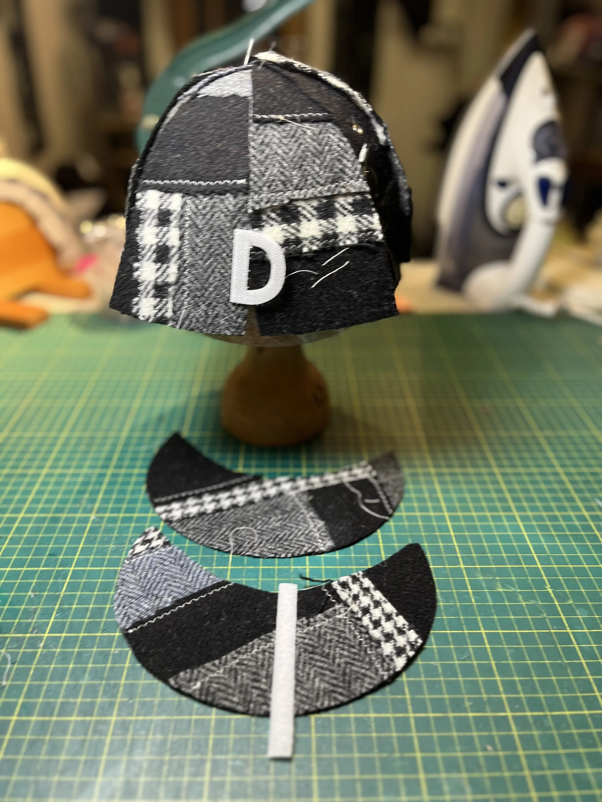 The creation of a bespoke Makshy cap. Panels of black-and-white houndstooth and check tweeds are pinned into a crown on a wooden block, featuring the custom initial 'D'. In the foreground, the matching brim pieces are ready for sewing.