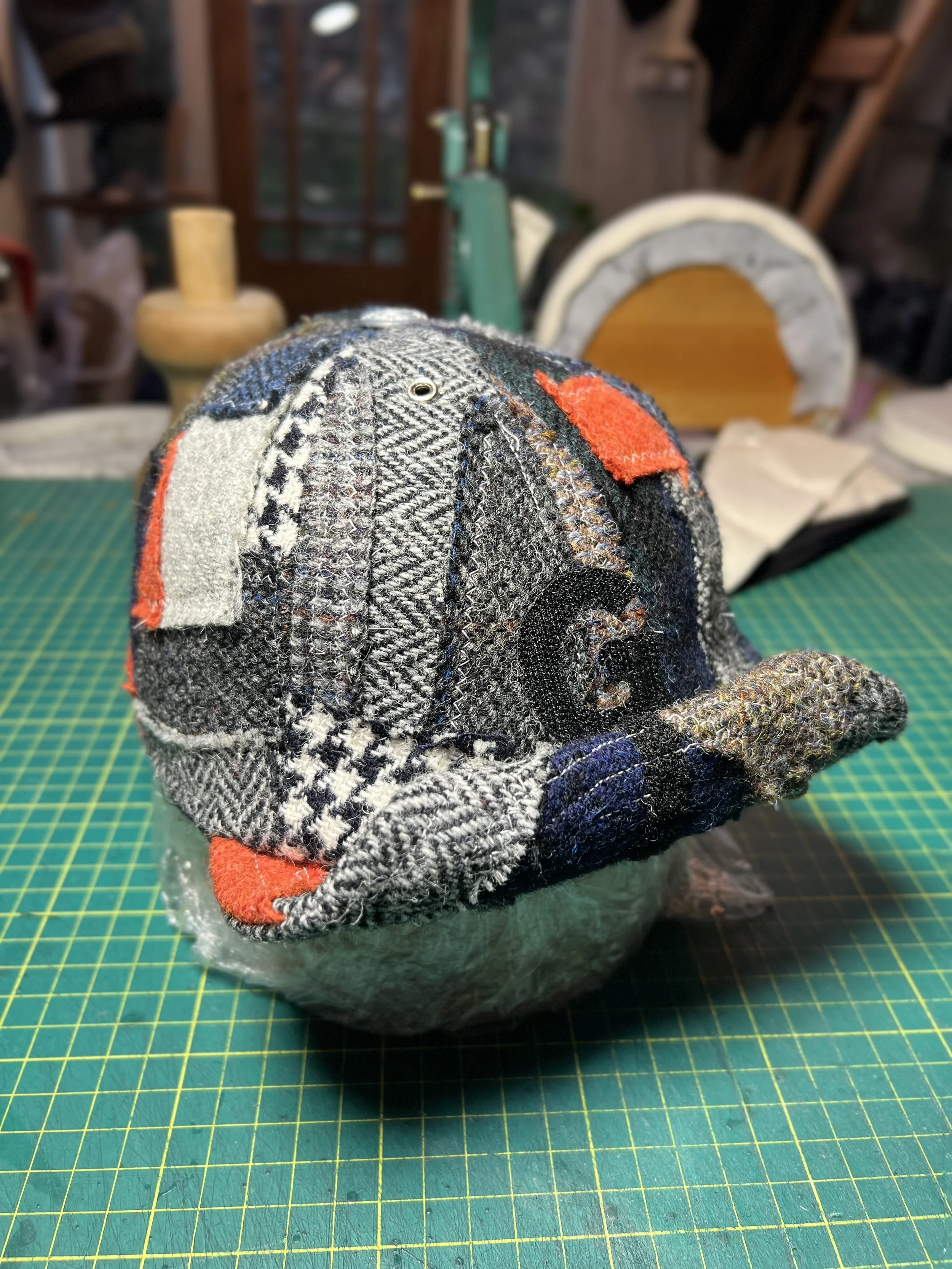 A one-of-a-kind patchwork cap by Makshy Millinery, displayed on a green cutting mat in the studio. The design features an intricate mix of traditional tweeds contrasted with bold orange panels.