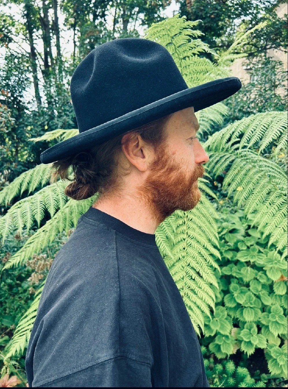 A man with reddish-brown hair and a beard, wearing a large, black Makshy fedora with a very wide, flat brim. He is shown in profile against a background of lush green ferns.