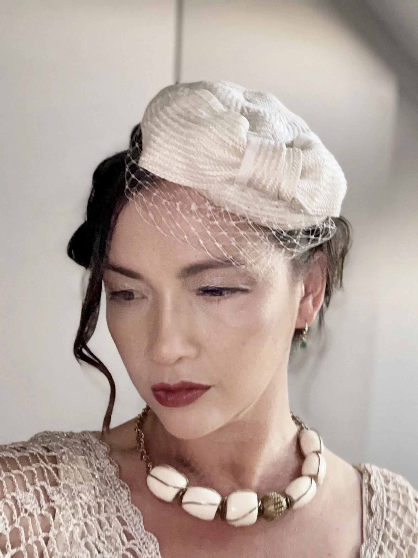 A woman with vintage-styled dark hair, wearing an off-white Makshy visca straw fascinator. The hat, featuring a decorative bow and a delicate birdcage veil, is paired with a chunky beaded necklace for a classic, elegant look.