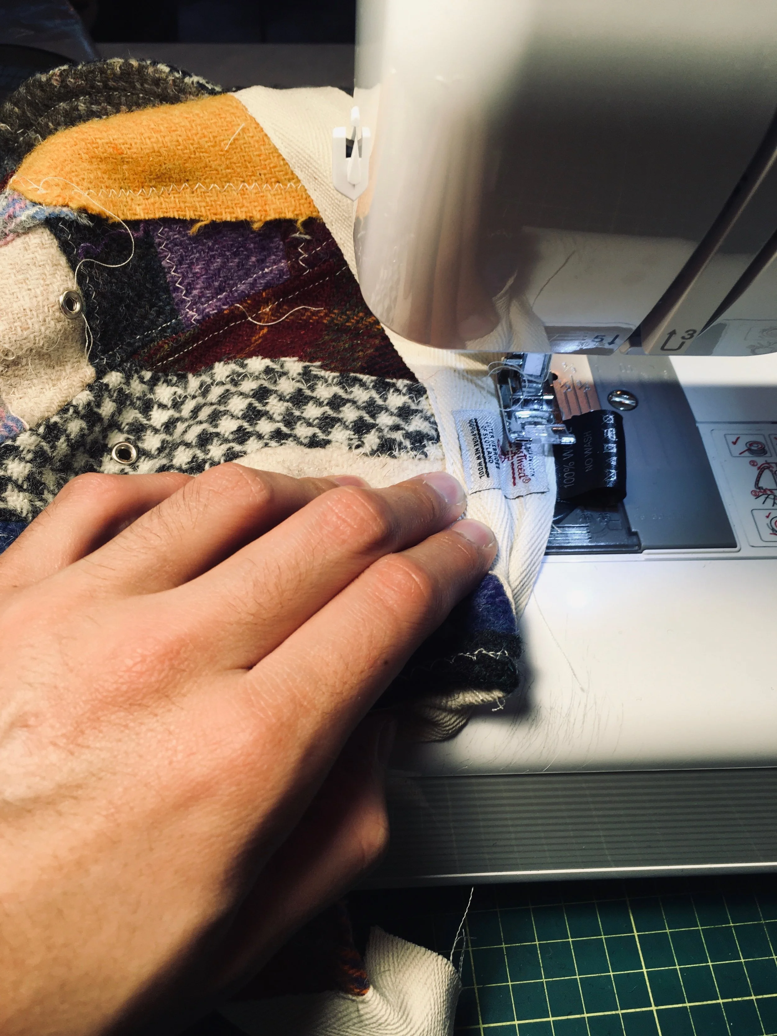 The hands-on creation of a Makshy Millinery piece. A milliner's hand guides a quilted patchwork of warm, multi-colored wool tweeds as it is stitched on a sewing machine.