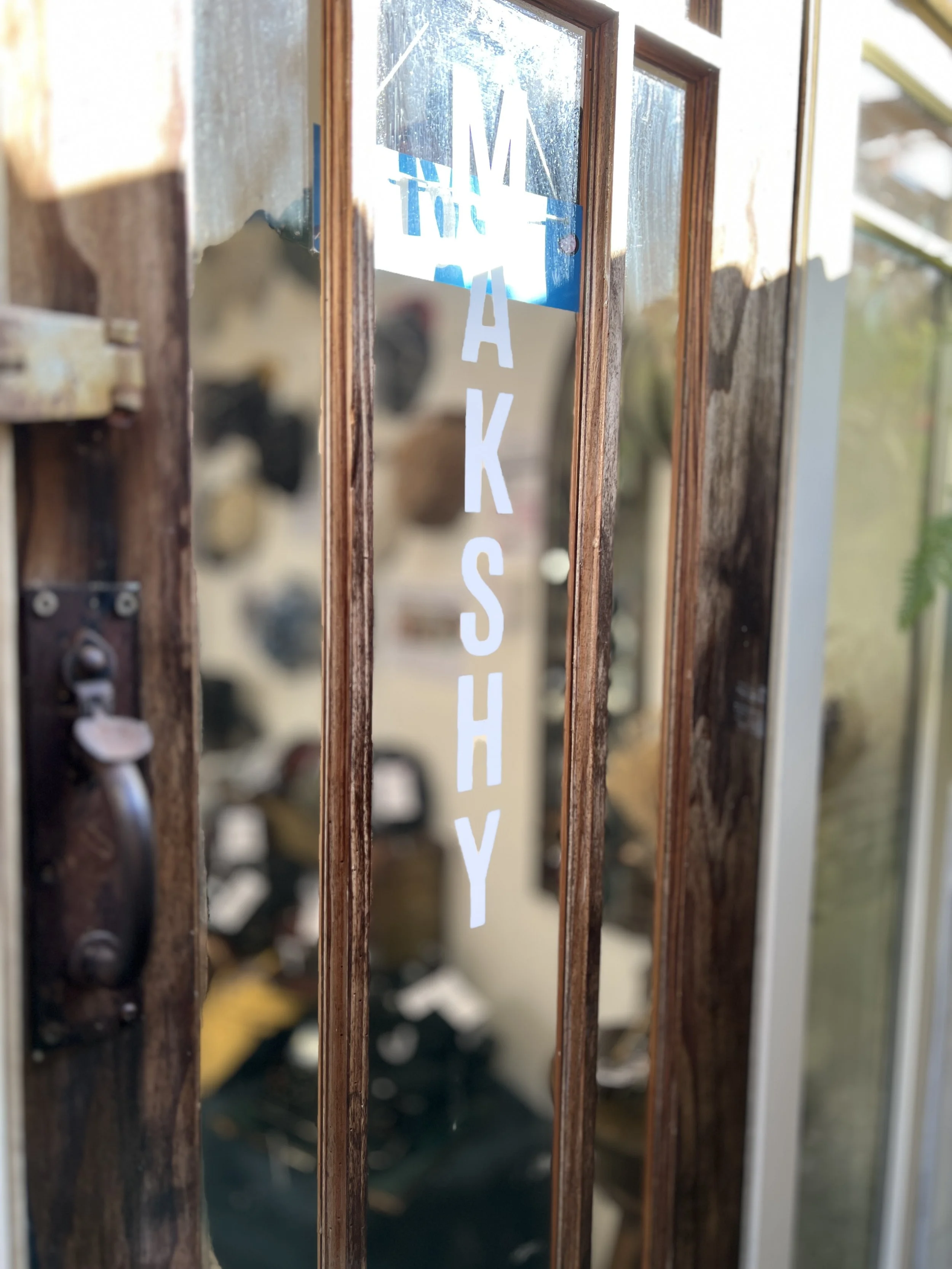 The entrance to the Makshy London atelier — a wooden door with the brand’s name etched on glass, opening into a workspace filled with handcrafted hats.