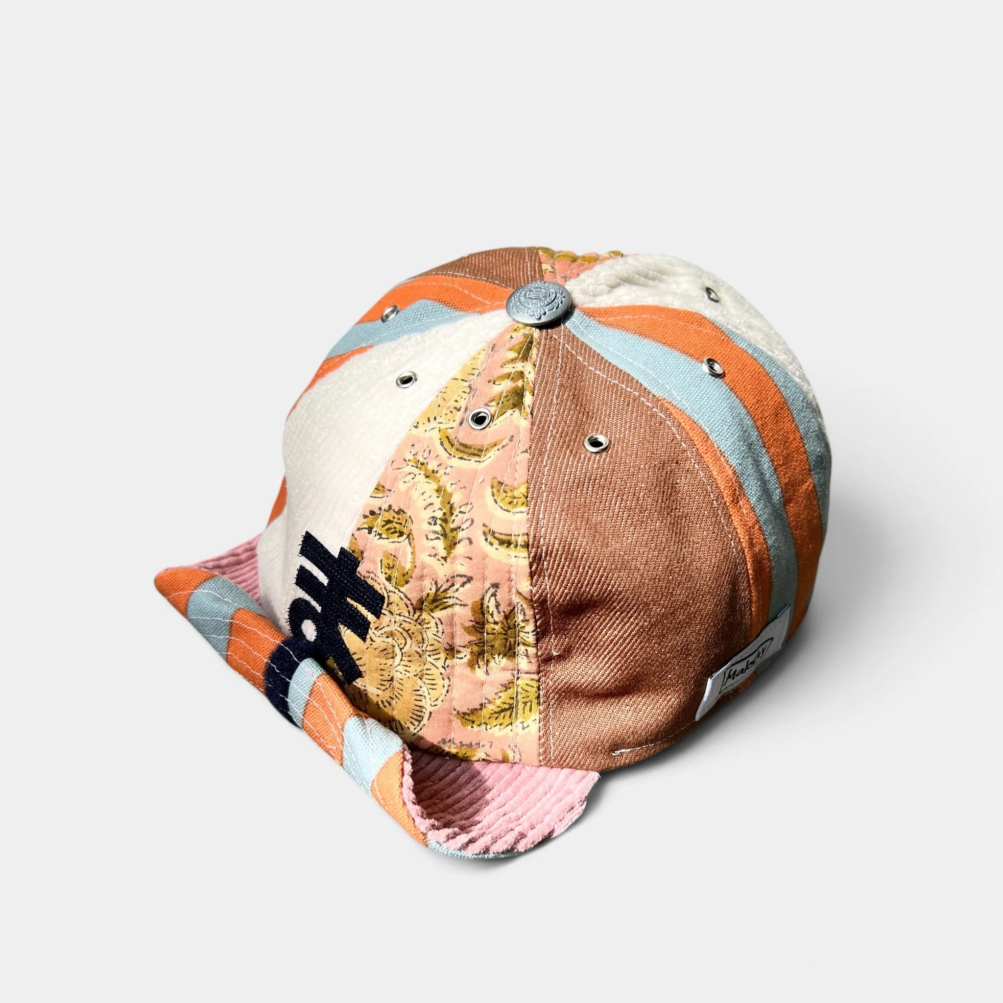 Mixed-panel cotton cap with roll-up visor, navy greeting motif, botanical-print panel and orange-and-turquoise striped accents.