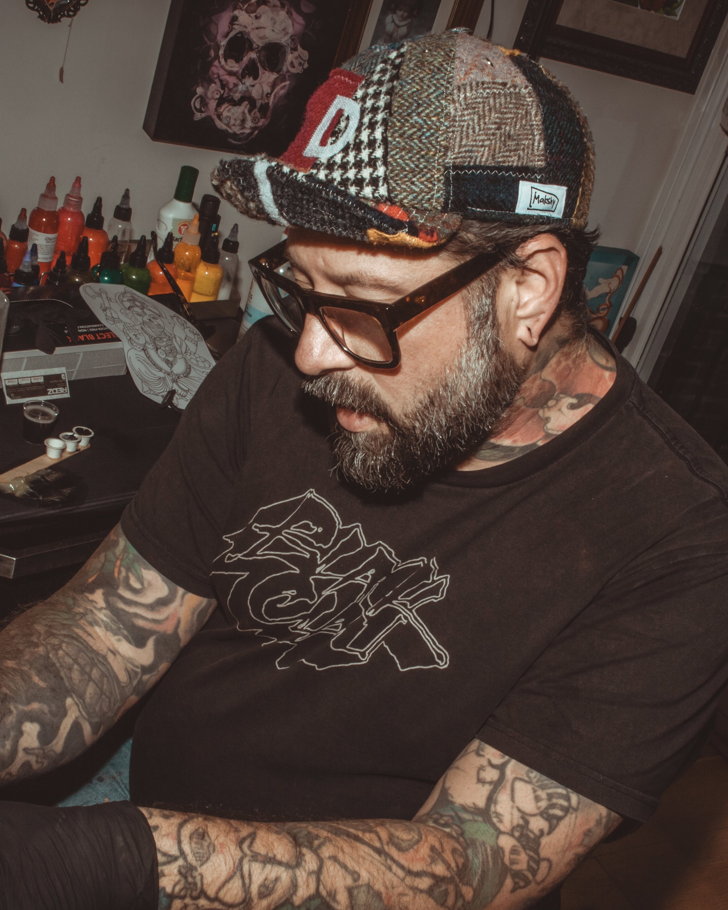 A man with tattoos, a beard, glasses, and a patchwork cap is sitting at a table in a tattoo studio with tattoo supplies and artwork around him.