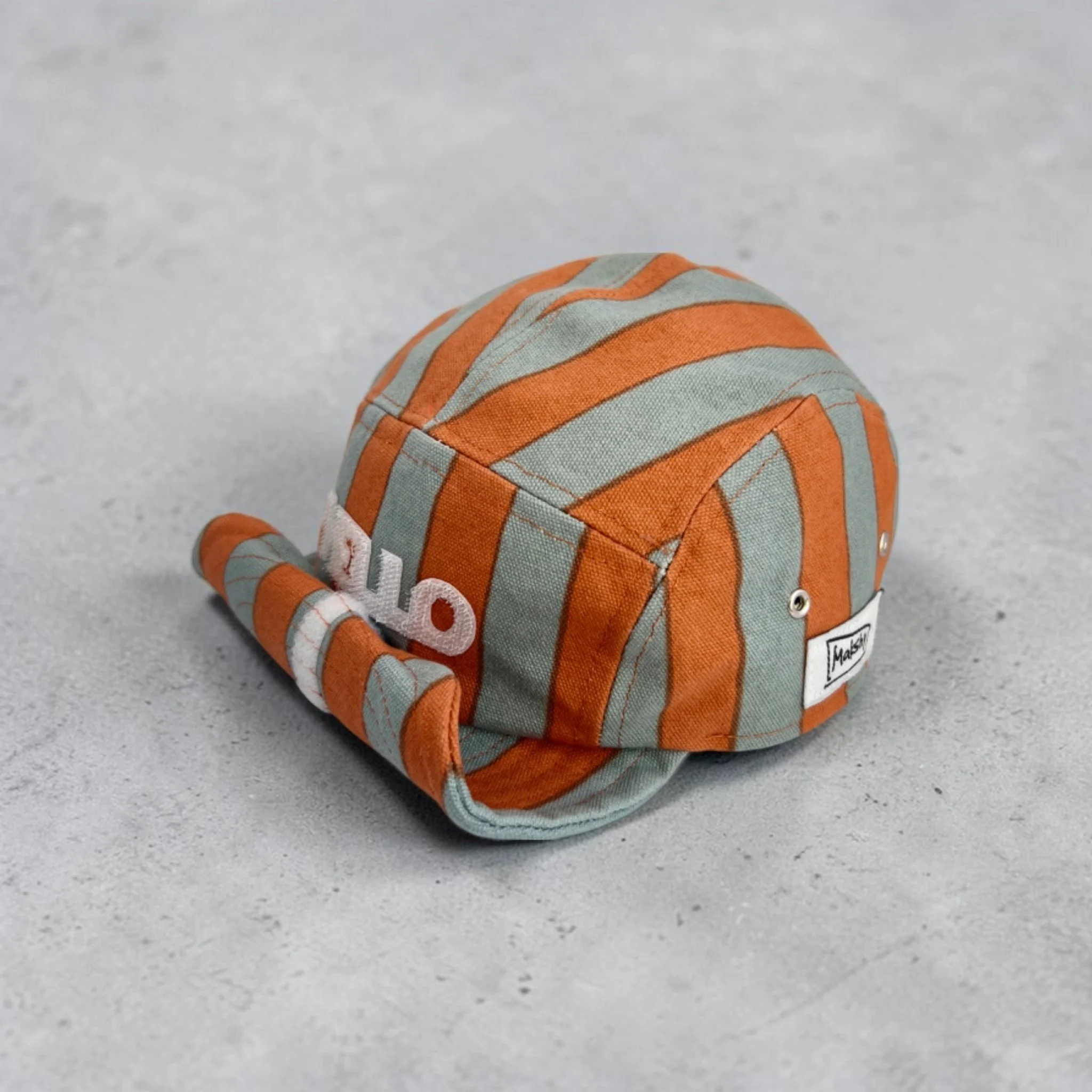 Orange and sage striped 5 panel cotton baseball cap with a structured crown, metal eyelets, and a shapeable roll-up visor, featuring a white velcro HELLO motif on the front panel, set against a neutral grey background.