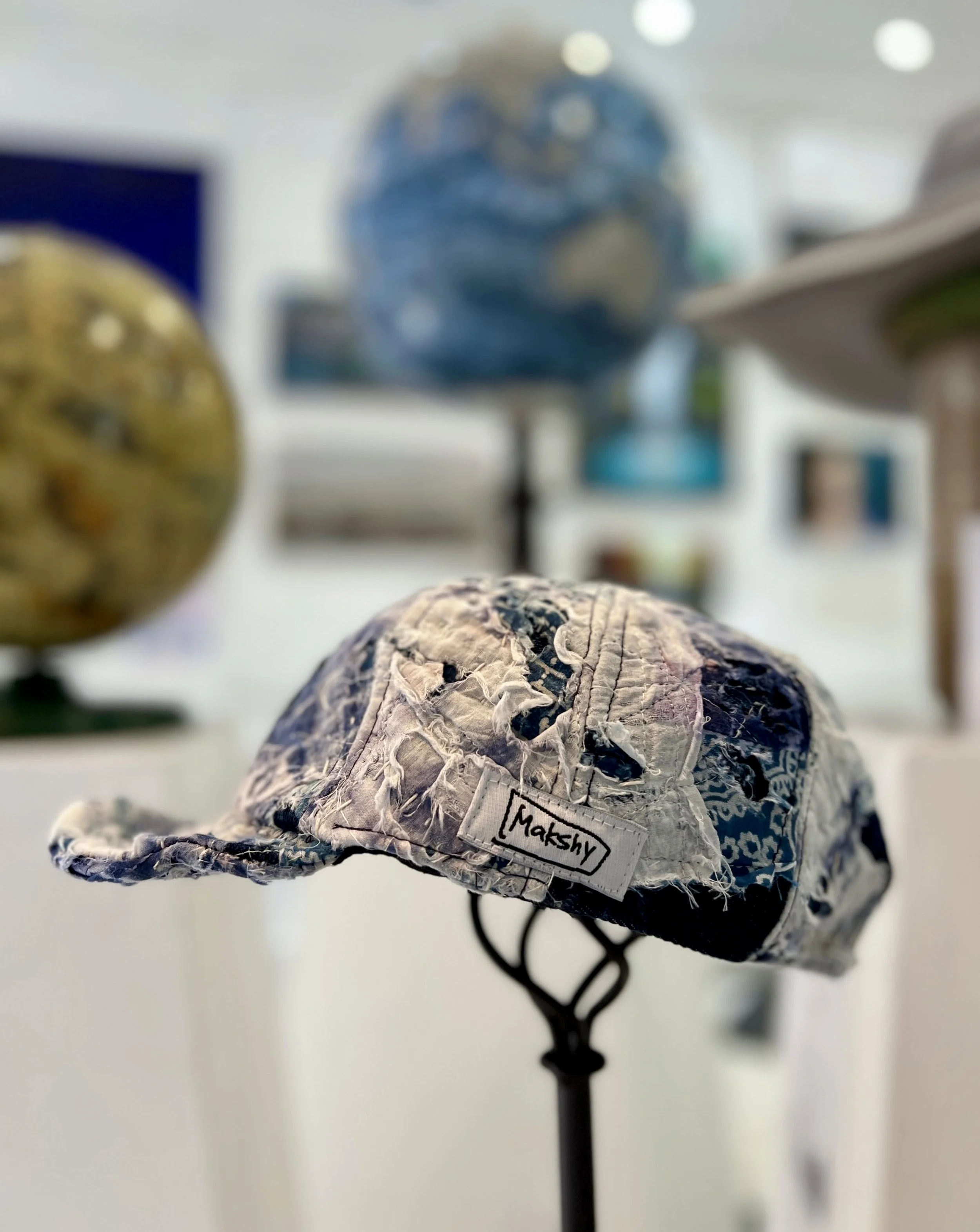 A Boro baseball cap with a label reading 'Makshy' displayed on a black stand, in an art gallery with various globes in the background.