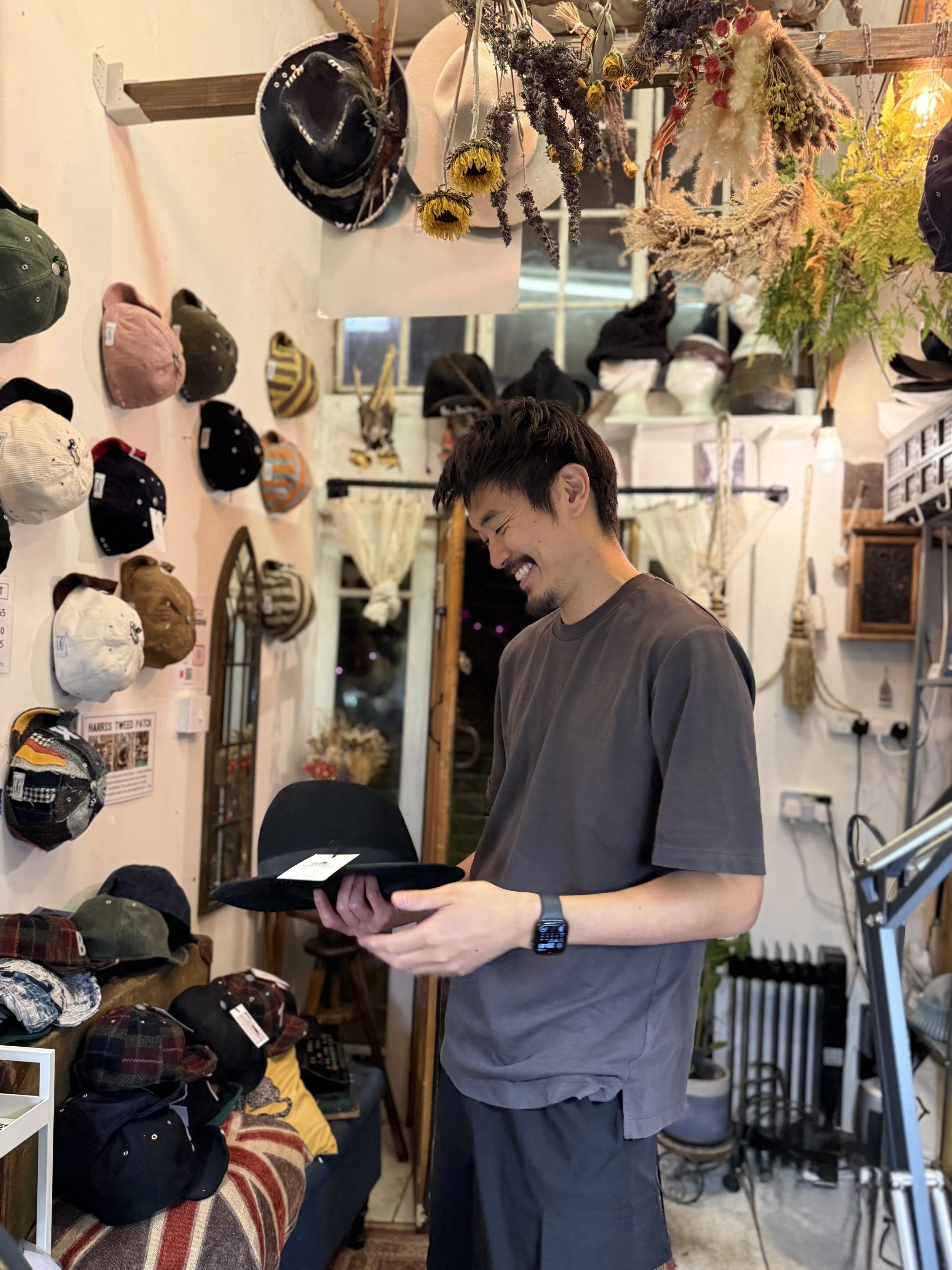 A young man with short dark hair and a beard, wearing a gray t-shirt, is smiling and looking at a black cap he is holding in a boutique hat shop. The shop has hats on the wall and shelves, with decorative dried flowers hanging overhead.