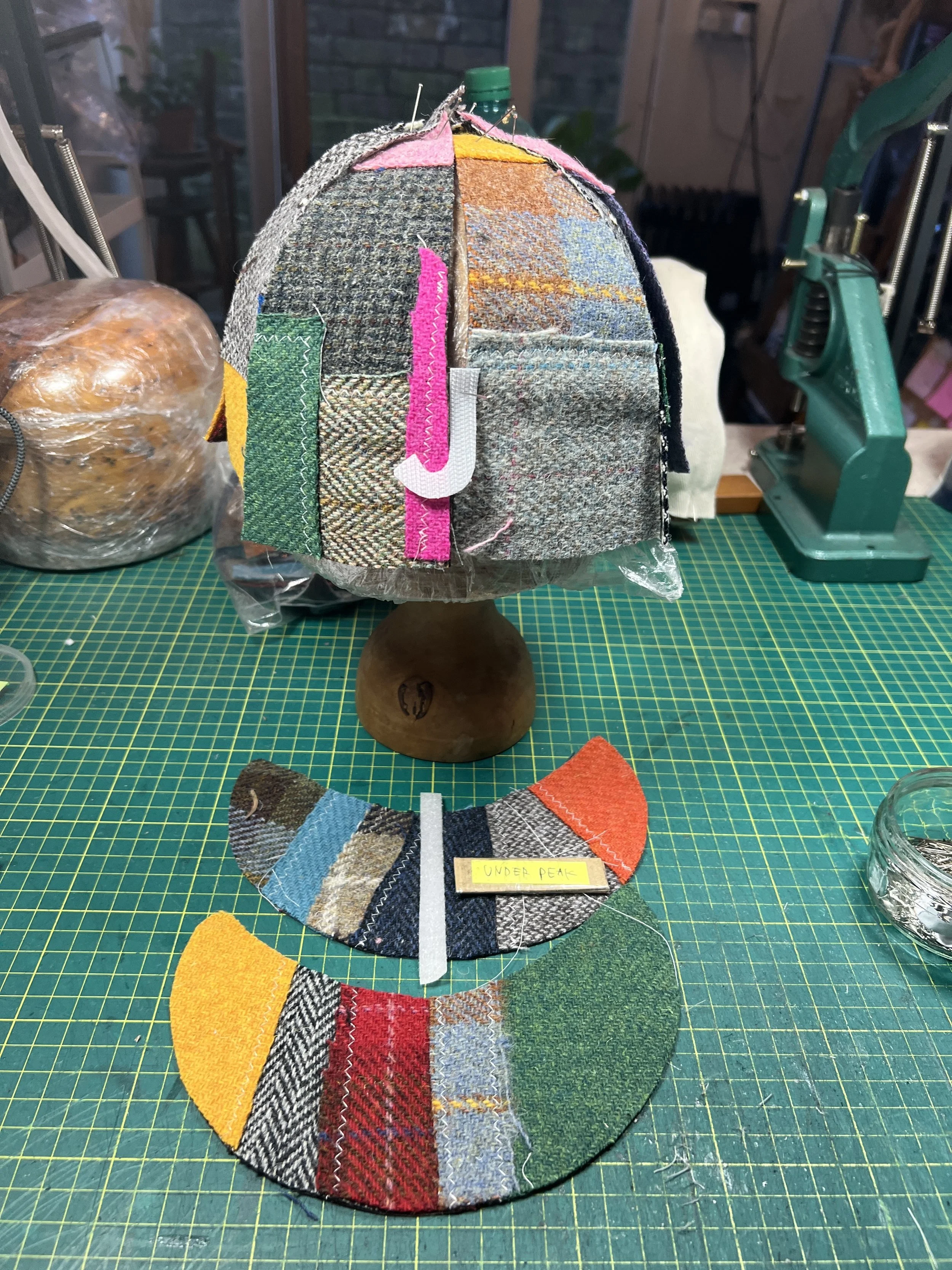 The creation process of a bespoke Makshy cap. A colorful Harris tweed patchwork crown is pinned into shape on a hat block, while the matching panels for the brim lie on the cutting mat, ready for the next step.