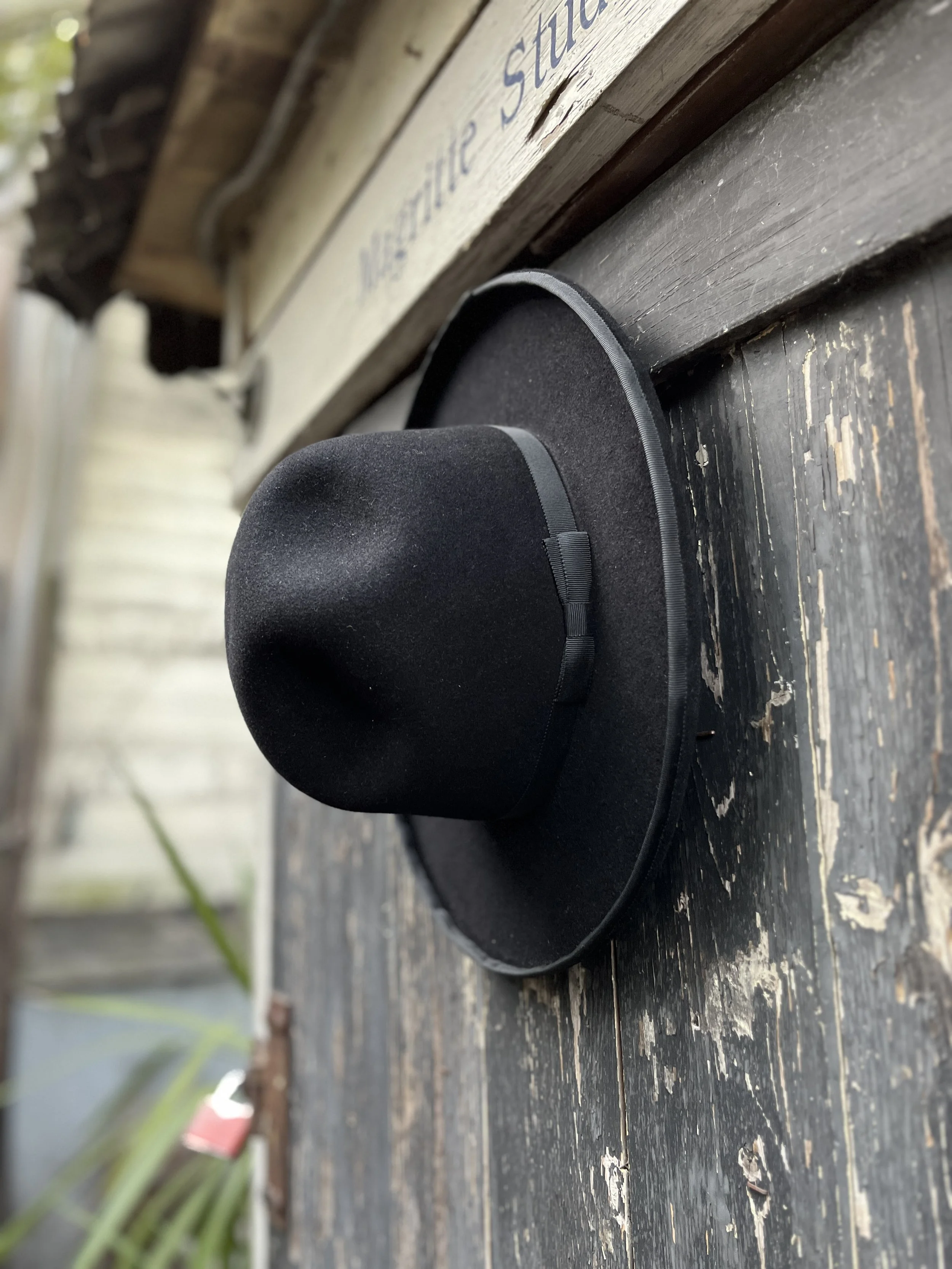 An atmospheric shot of a Makshy wool fedora, set against the richly textured surface of a peeling black wooden wall. The image evokes a rustic, artistic feel, like a hat hanging outside a creative studio.