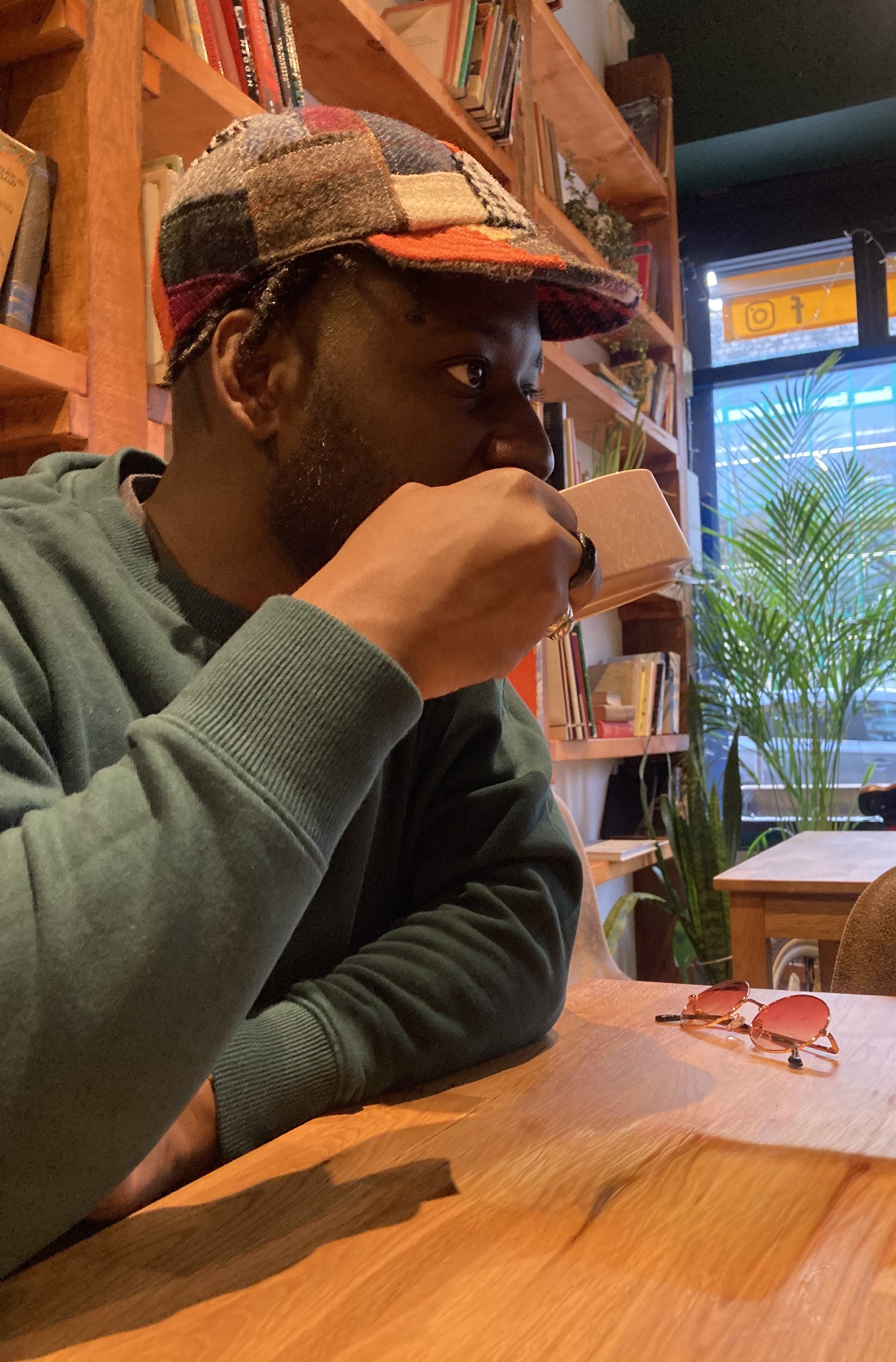 A candid lifestyle shot of a man enjoying a quiet moment in a cafe while wearing a unique Makshy patchwork cap. The warm, book-lined setting complements the classic, artisanal style of the tweed hat.