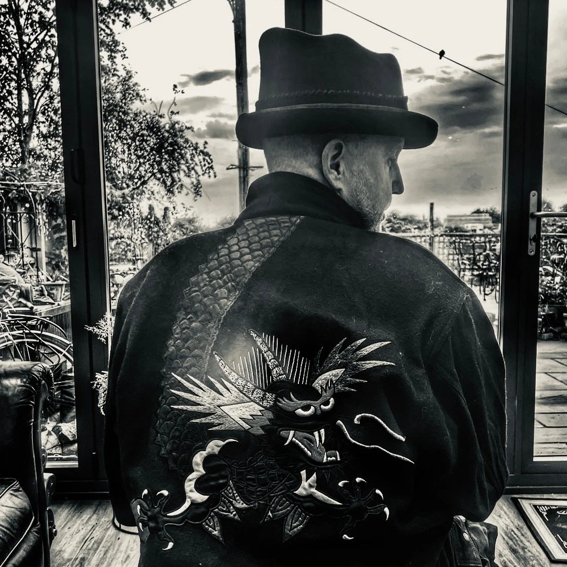 A stylish man, seen from behind, wears a Makshy Mohican Rhombus Fedora while looking out a window. His dark jacket features a large, embroidered dragon on the back. The atmospheric photo is in black and white.