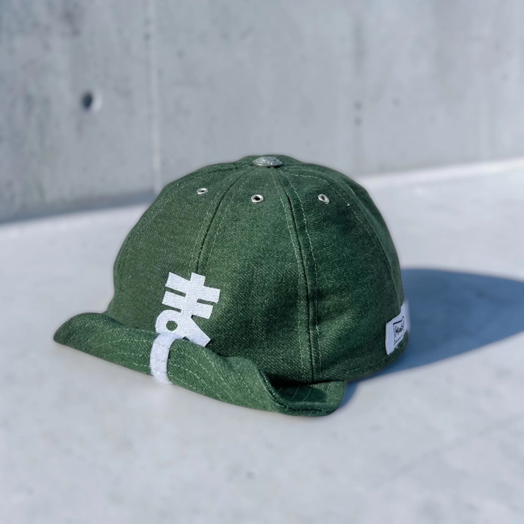 Green corduroy 8 panel baseball cap with a structured crown, metal eyelets, and a shapeable roll-up visor, featuring a white hiragana ま (ma) motif on the side, set against a neutral grey background.