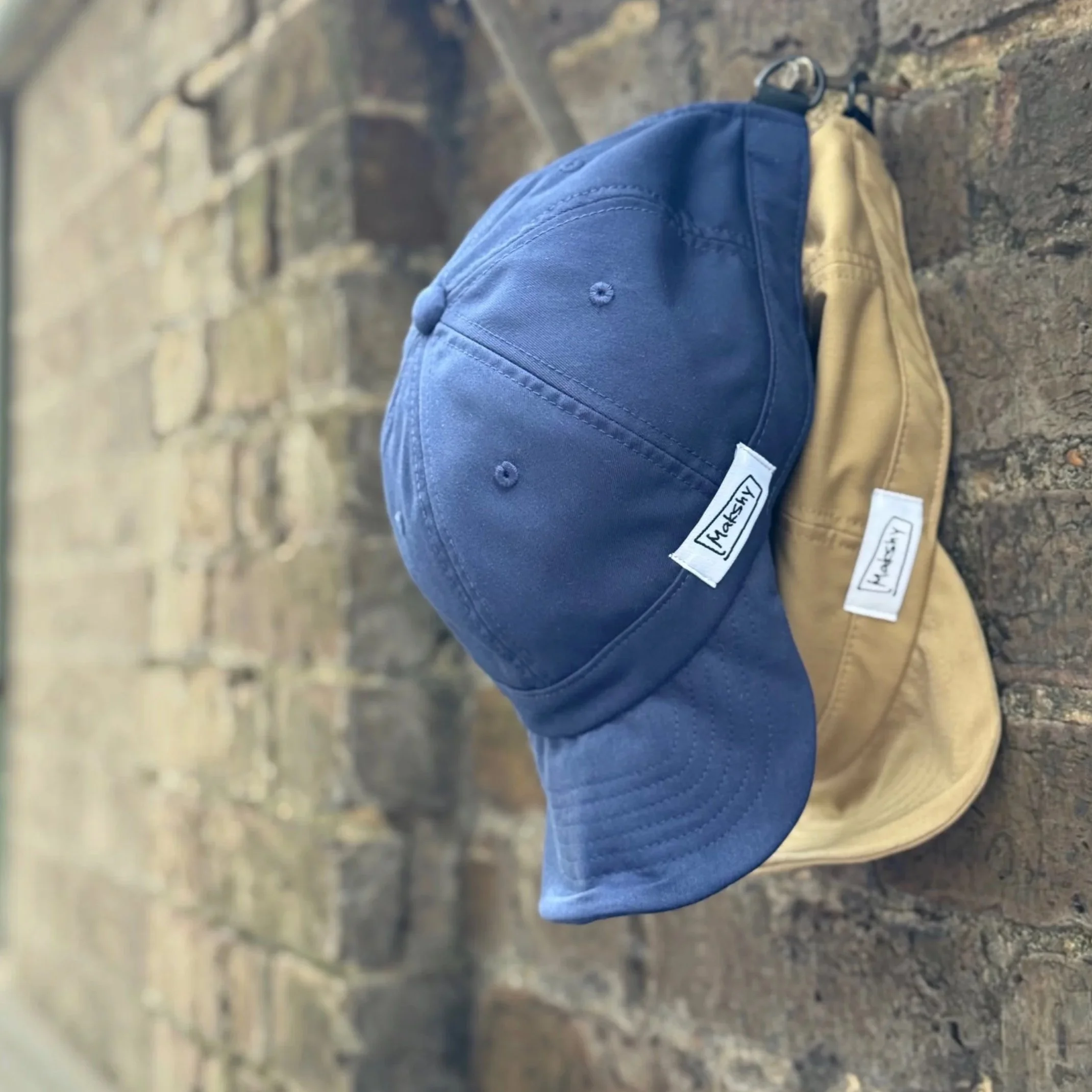 Navy and beige six-panel baseball caps with shapeable wired visors and D-ring loops hanging from a brick wall.