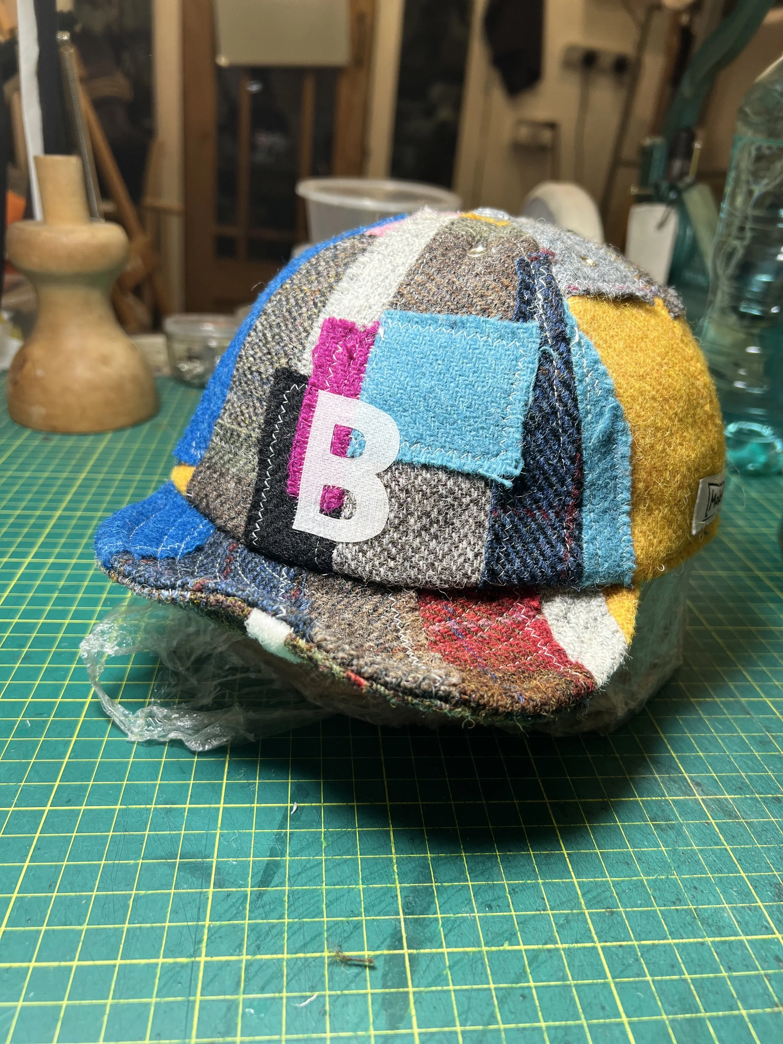 A bespoke, multi-colored patchwork cap from Makshy Millinery, displayed on a studio workbench. This one-of-a-kind piece is made from a rich assortment of colorful tweeds and is personalized with the initial 'B' on a bright blue patch.