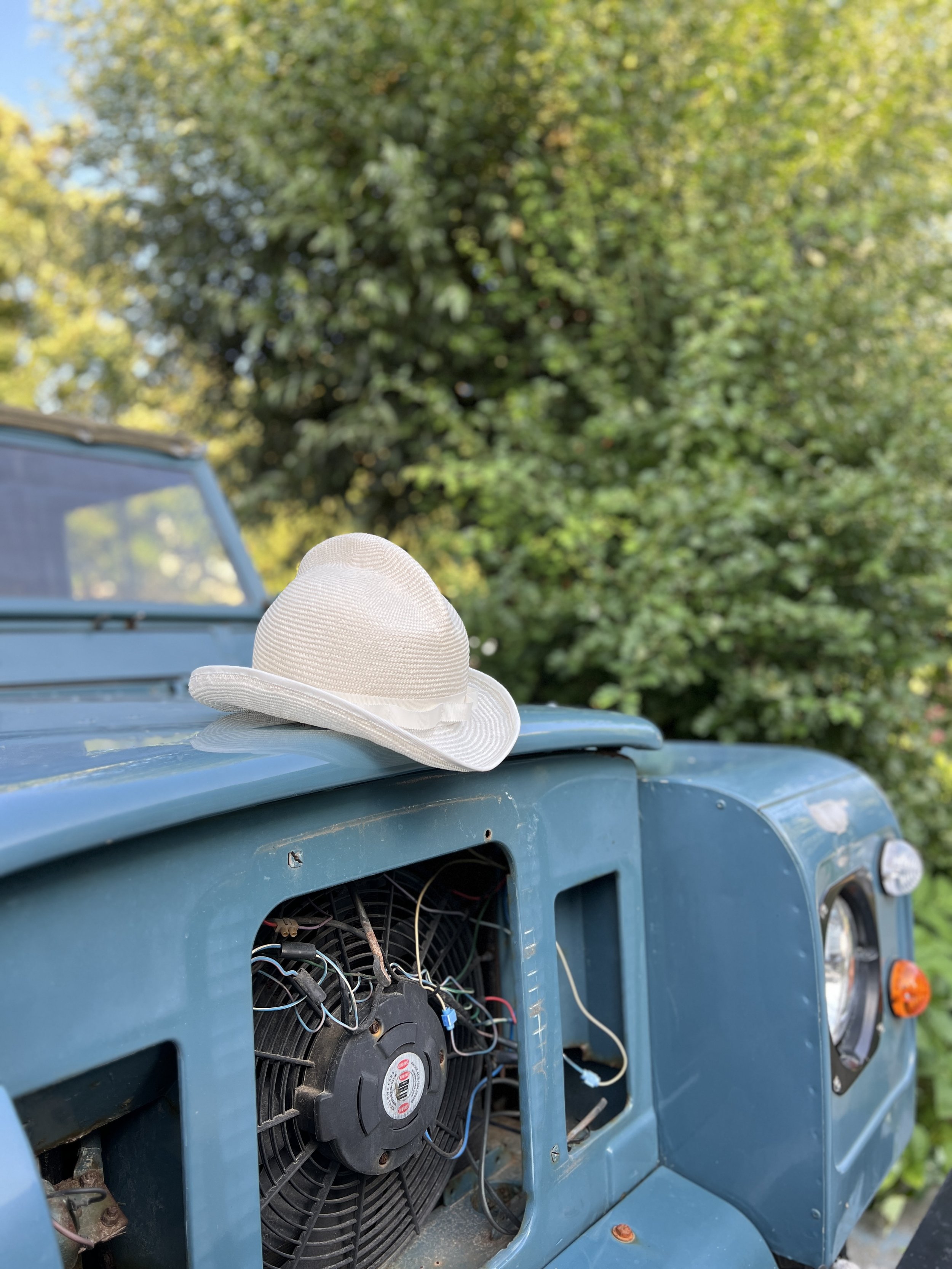 A stylish, off-white Makshy visca straw hat resting on the blue bonnet of a vintage Land Rover Defender. The vehicle's grille is removed, showing the engine fan, and the background is a lush green garden.
